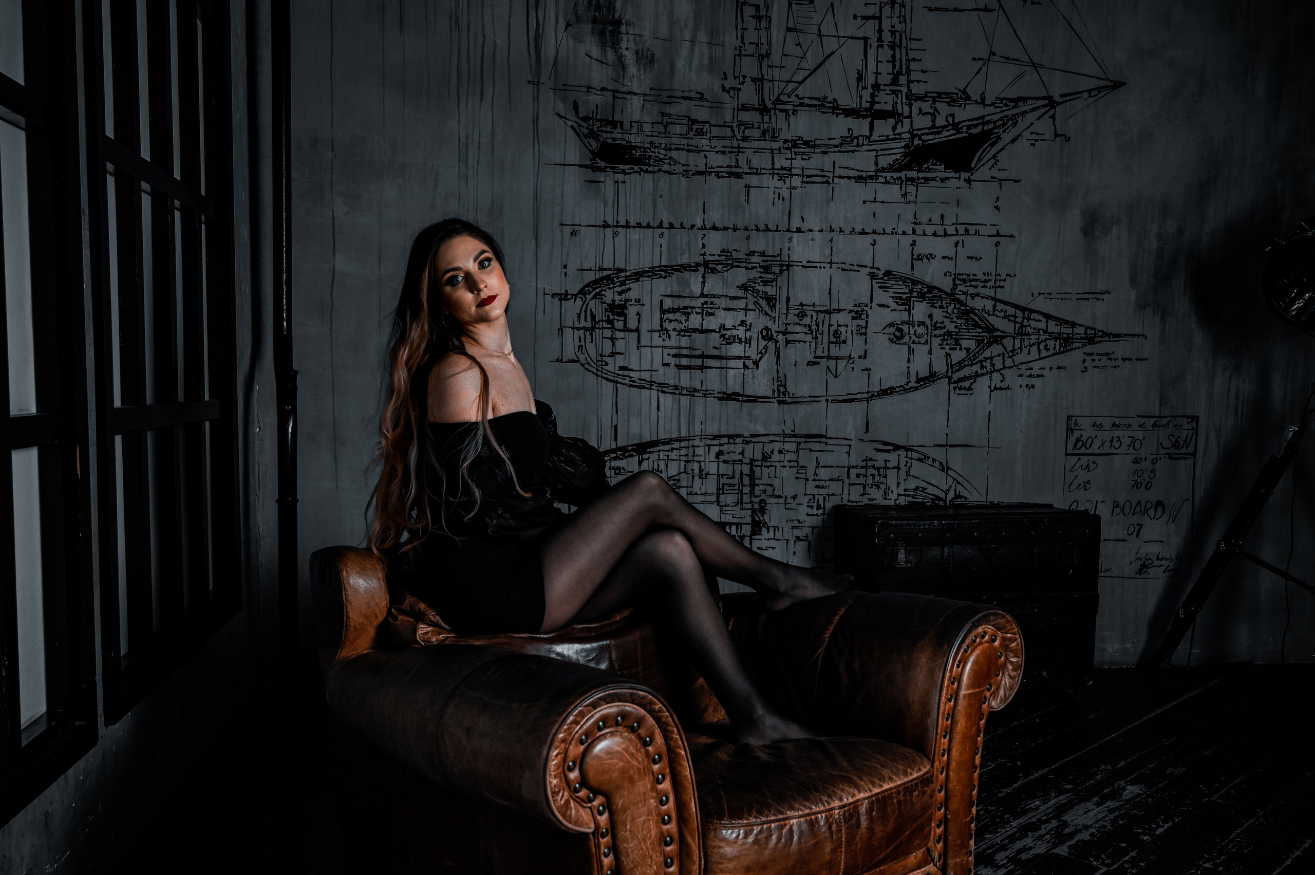 Молодая девушка сидит на кожаном диване в темной комнате. Она одета в черное платье и чулки, демонстрируя элегантный и загадочный образ. Ее длинные волосы свободно падают по плечам, а взгляд направлен прямо в камеру, создавая ощущение интимности и сосредоточенности. На стене за ней видны черно-белые технические рисунки кораблей и документация, что придает помещению атмосферу таинственности и историчности. Освещение мягкое и направленное, акцентирующее внимание на модели и детали интерьера. Фотография передает атмосферу мистики и утонченного стиля.