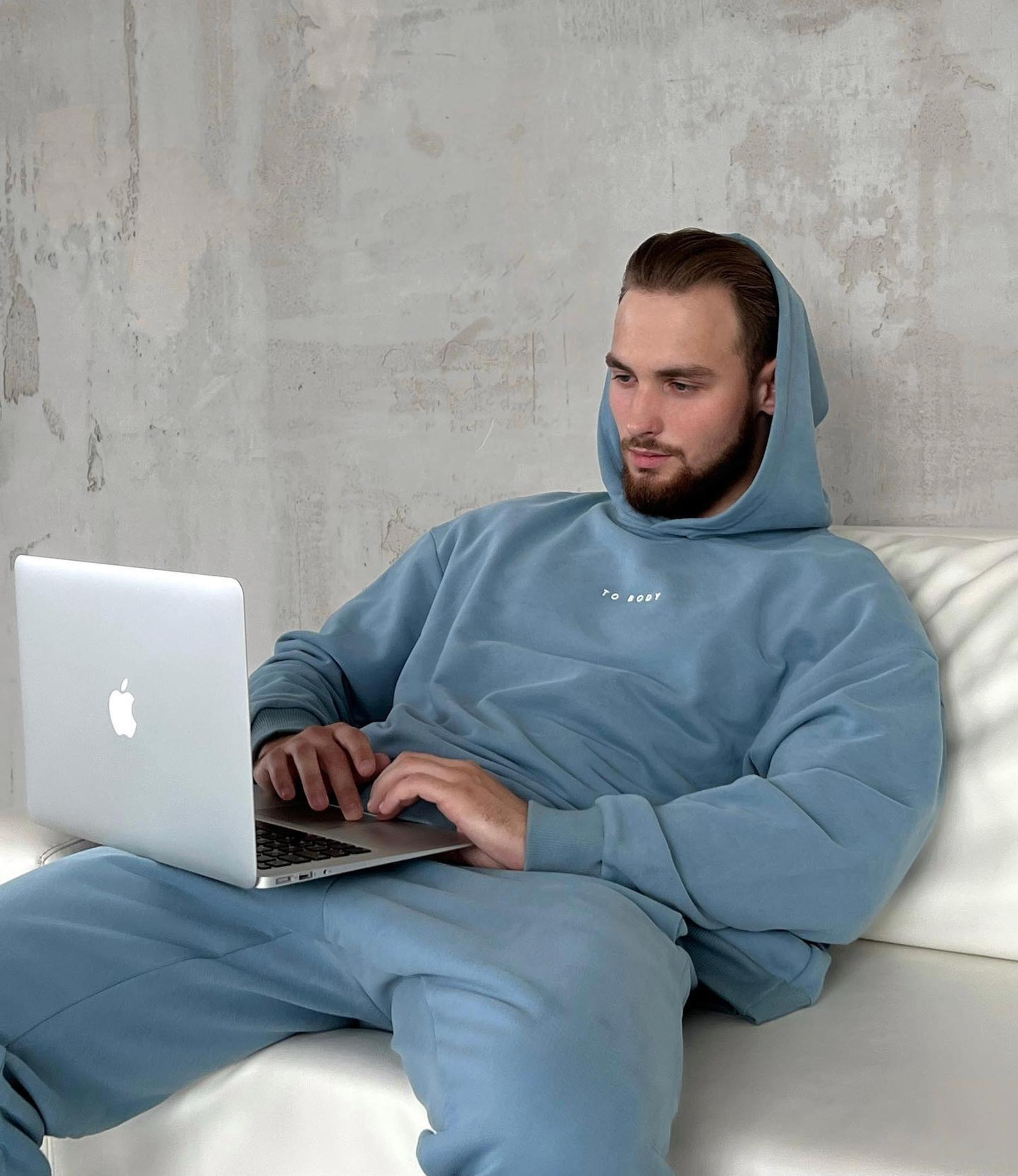 Мужчина с бородой в голубом худи сидит на белом диване с ноутбуком на коленях