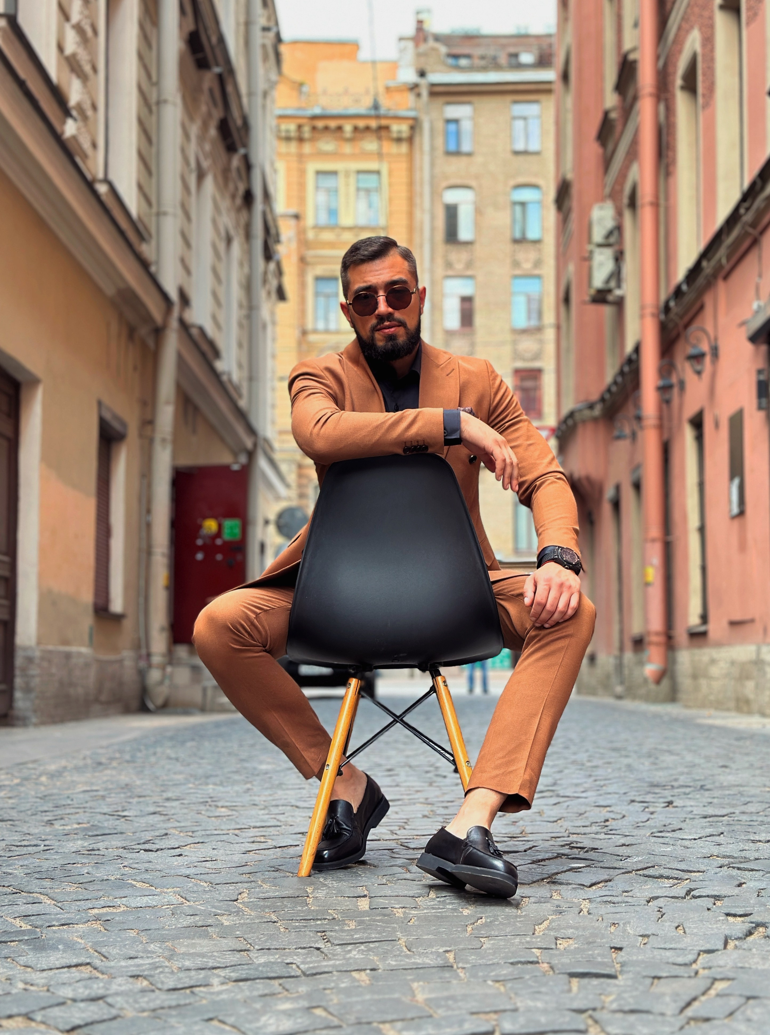Мужчина с бородой в оранжевом костюме сидит на стуле посреди улицы в Питере