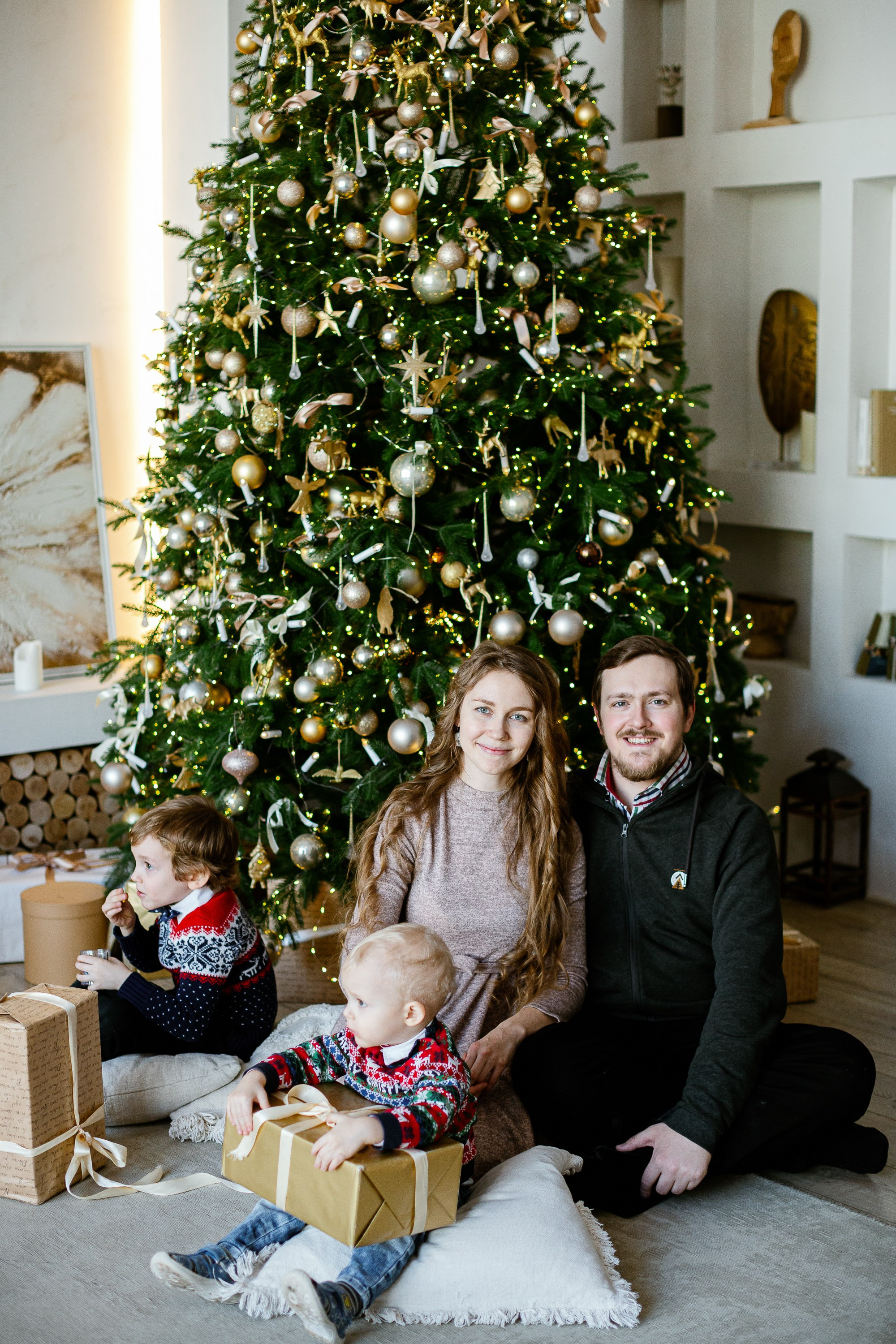 Семейная сидит на полу на фоне стоит елка и светится огоньками. Все улыбаются и смеются.