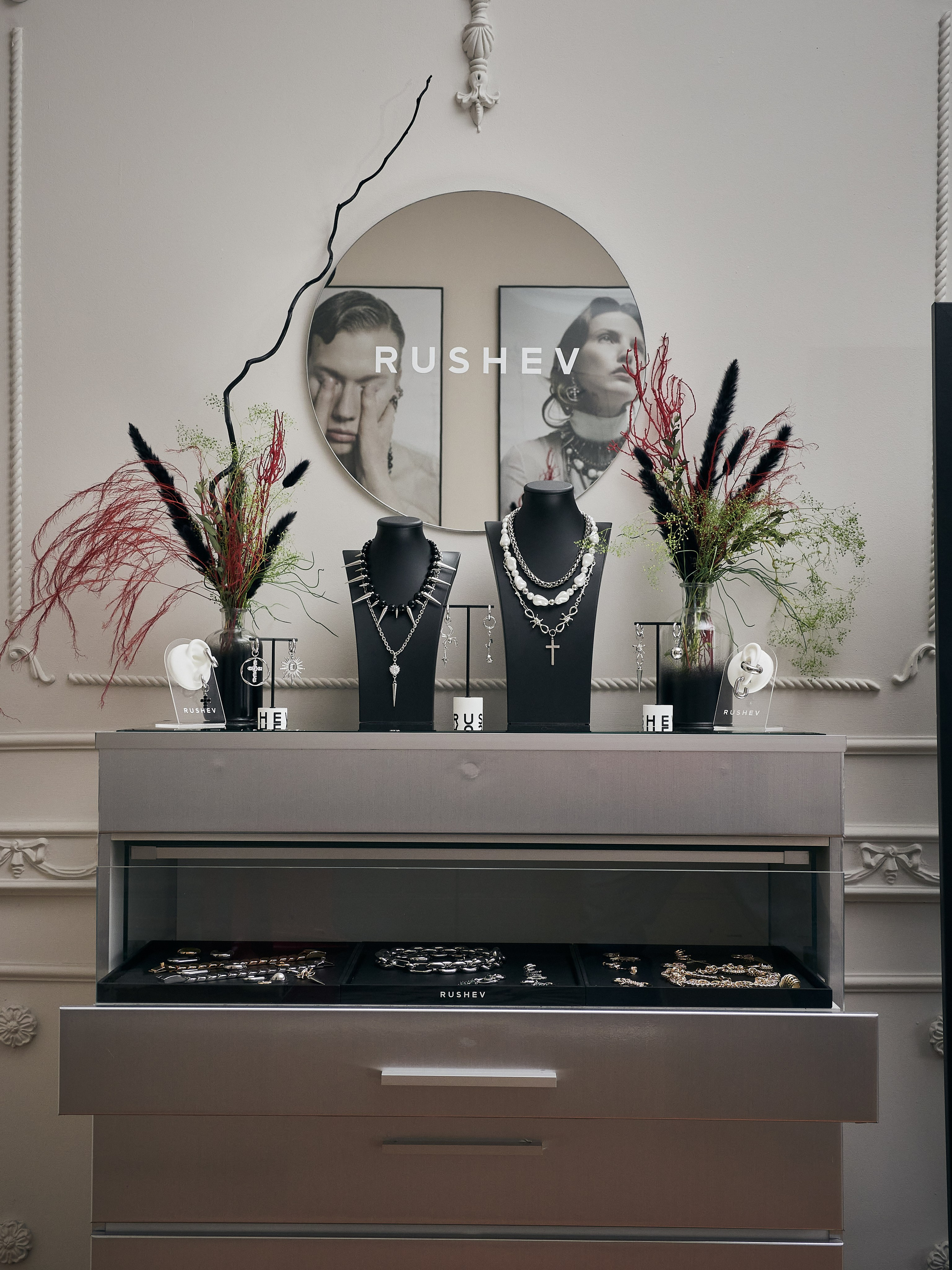 Элегантная витрина ювелирных изделий с ожерельями на подставках, декоративными растениями и зеркалом на стене с надписью RUSHEV