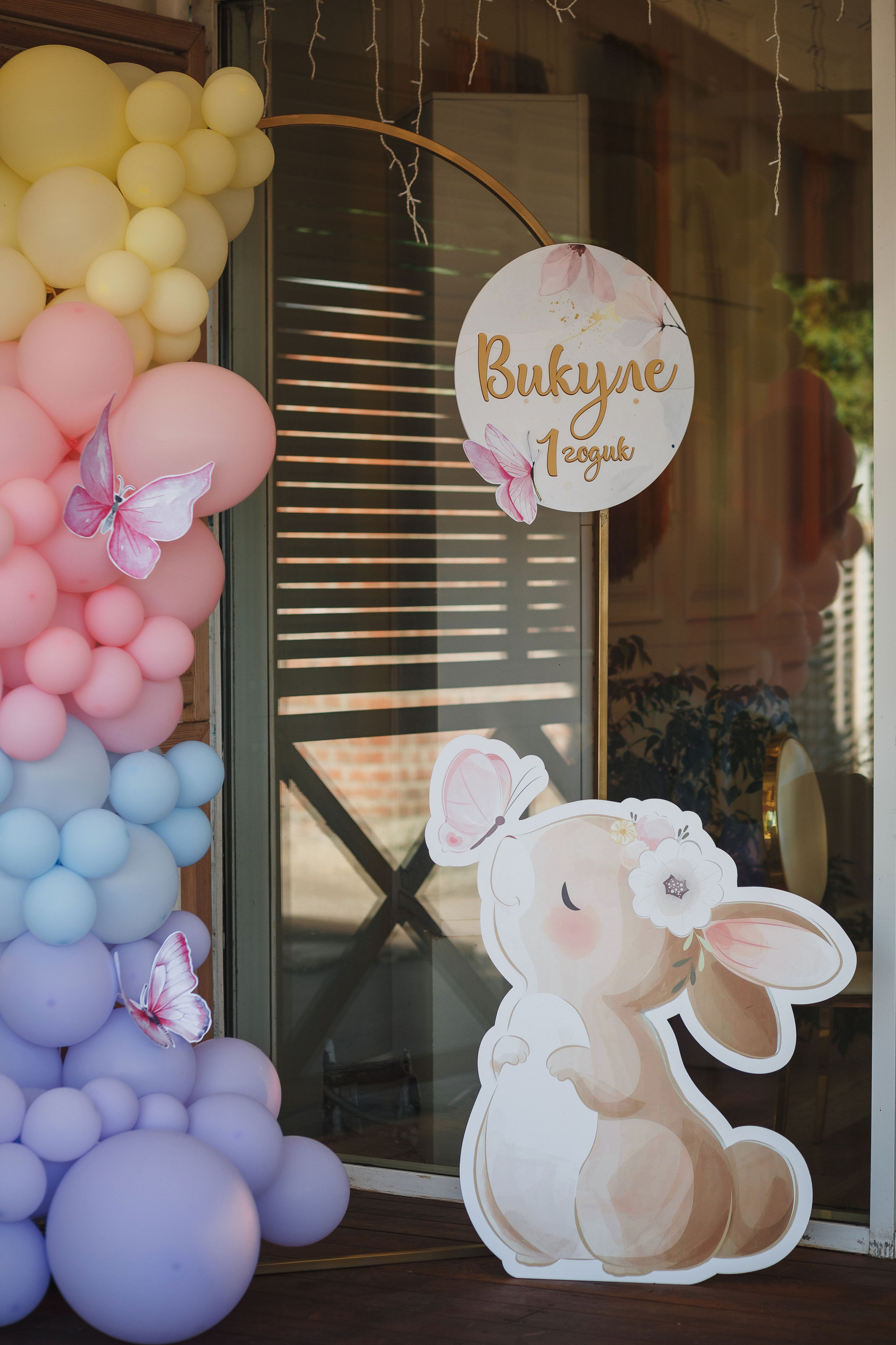 Фотозона для детского праздника с надписью 'Викуле 1 годик', украшенная воздушными шарами пастельных оттенков и милым рисунком зайчика с цветком. Фотозона расположена в Таганроге.