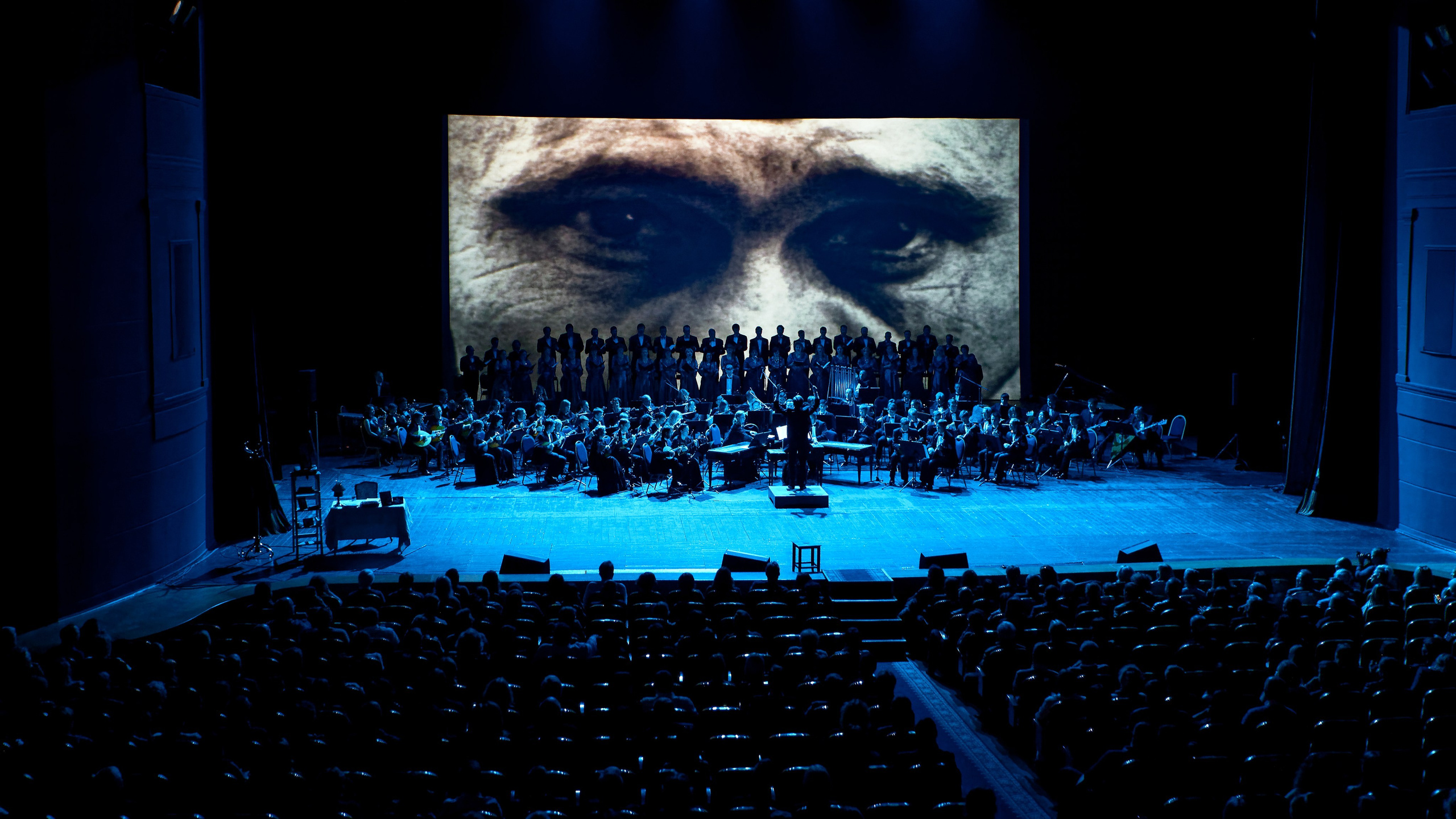 Масштабный концерт - большой симфонический оркестр и хор на сцене с огромной проекцией глаз на экране, синий сценический свет, полный зал зрителей