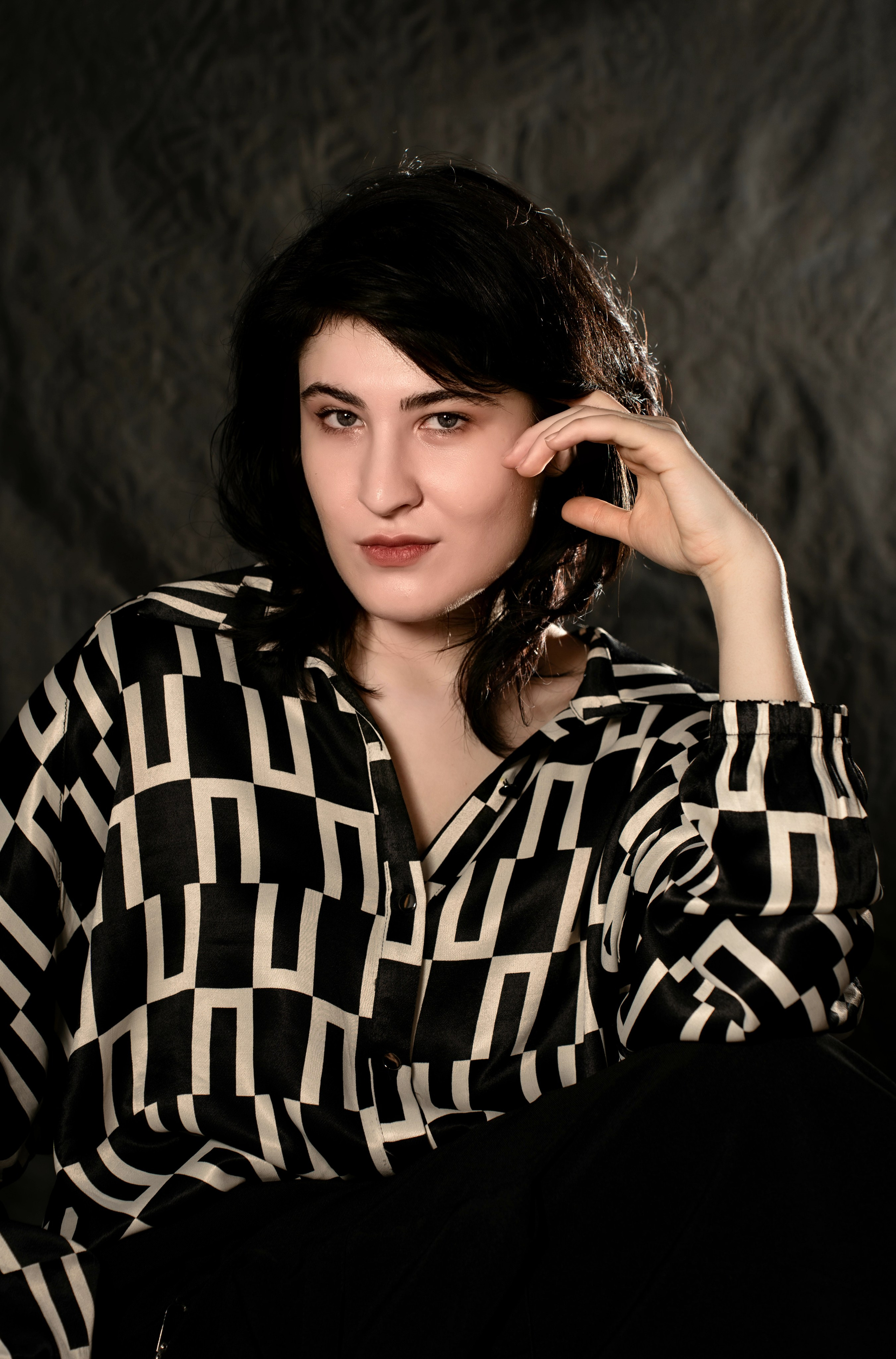 На изображении изображен человек с короткими темными волосами, одетый в черно-белую геометрическую блузку. Он сидит и позирует, одной рукой прижимаясь к лицу, сосредоточенно глядя в камеру. Фон текстурированный и темный, вероятно, это фон студии. 