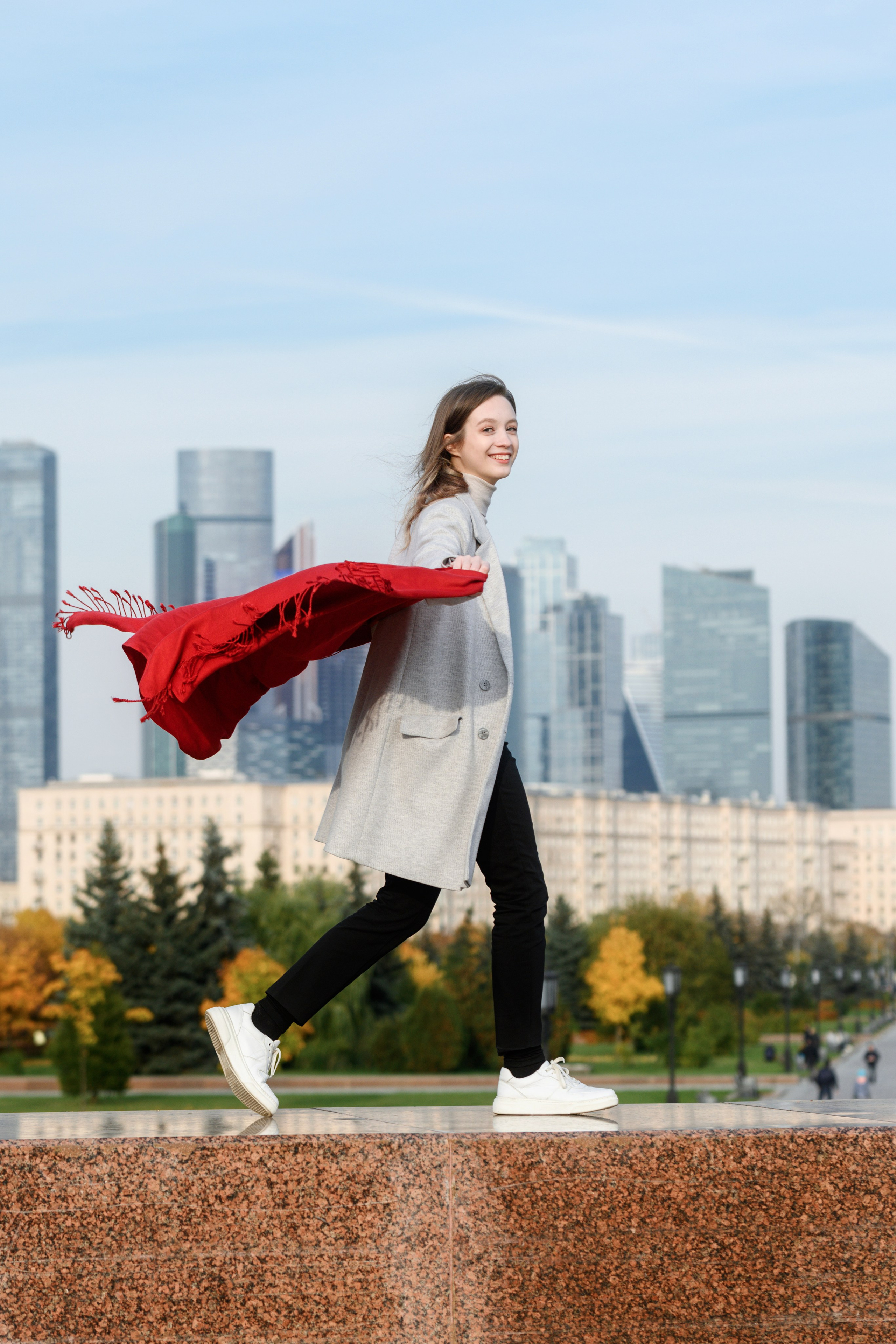 Девушка идёт с развевающимся красным шарфом на фоне городских высоток.