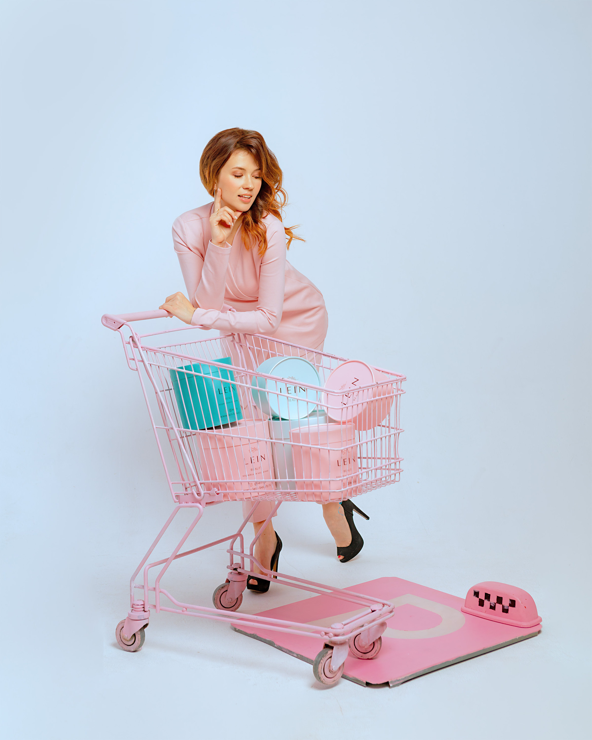 Девушка в розовом костюме стоит оперевшись на магазинную тележку наполненую коробками
