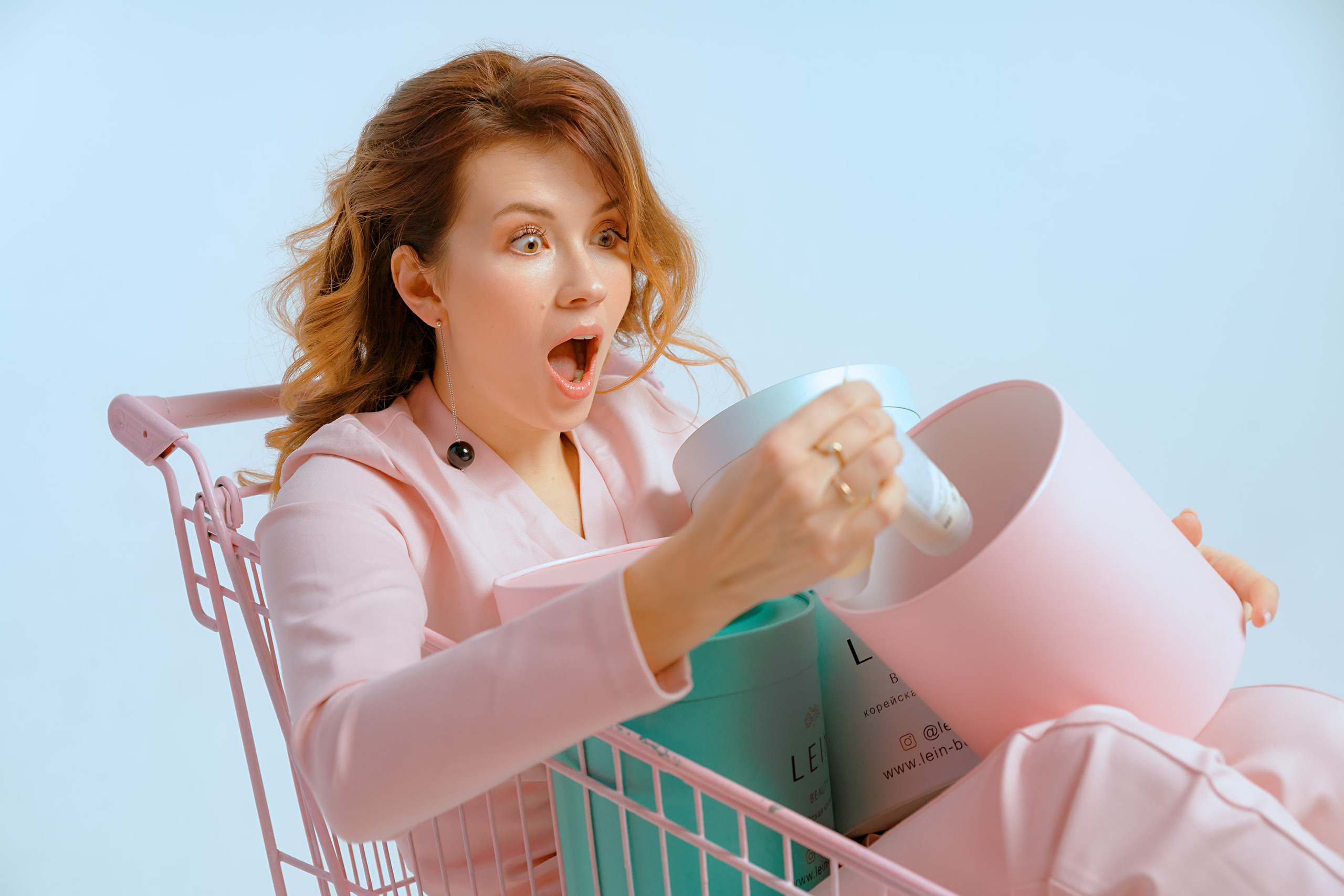 Девушка в розовом костюме с удивлением раскрывает коробку с косметикой сидя в магазинной тележке