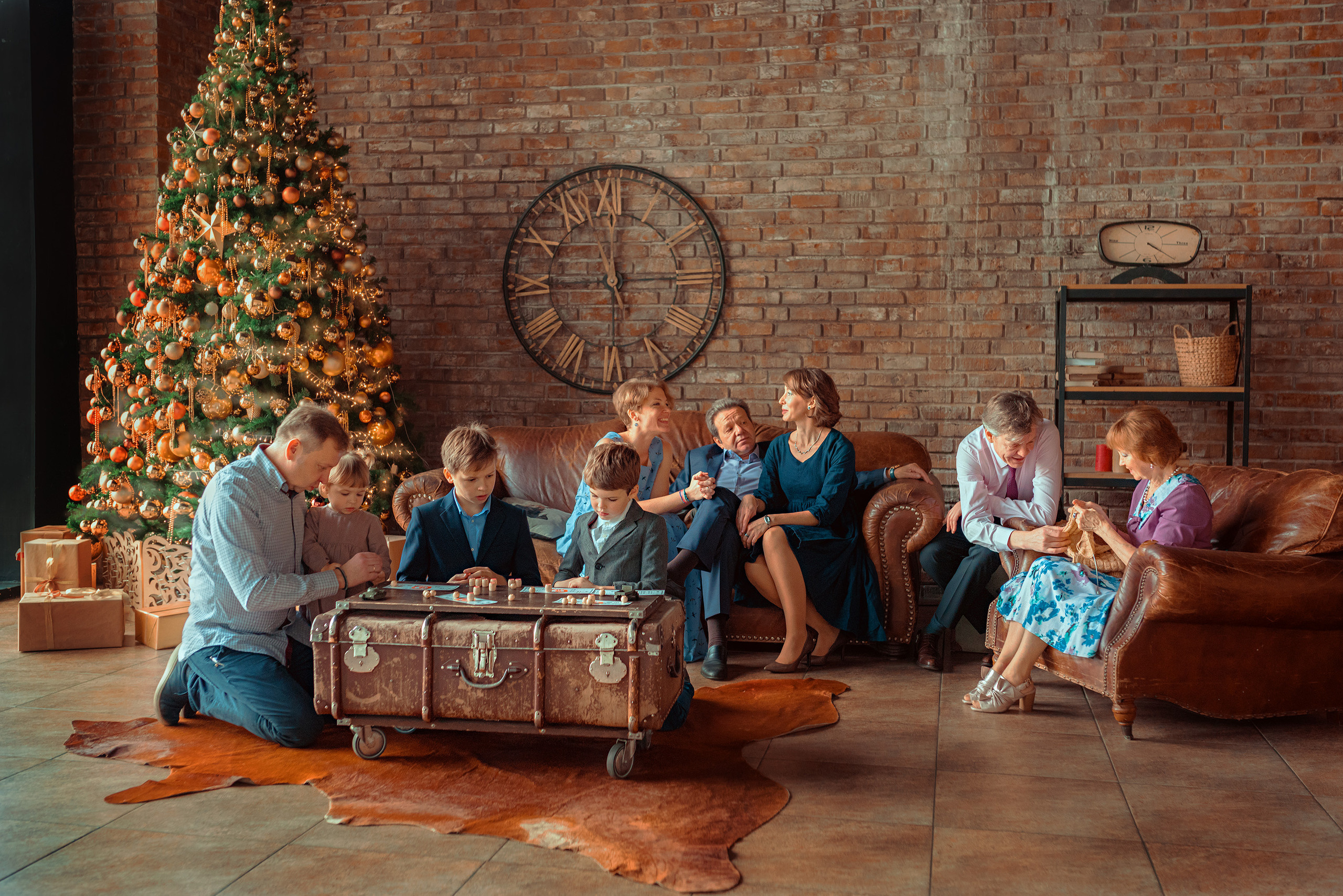 Большая семья общается сидя на диванах , часть из них играют в лото, на заднем плане большая наряженая елка