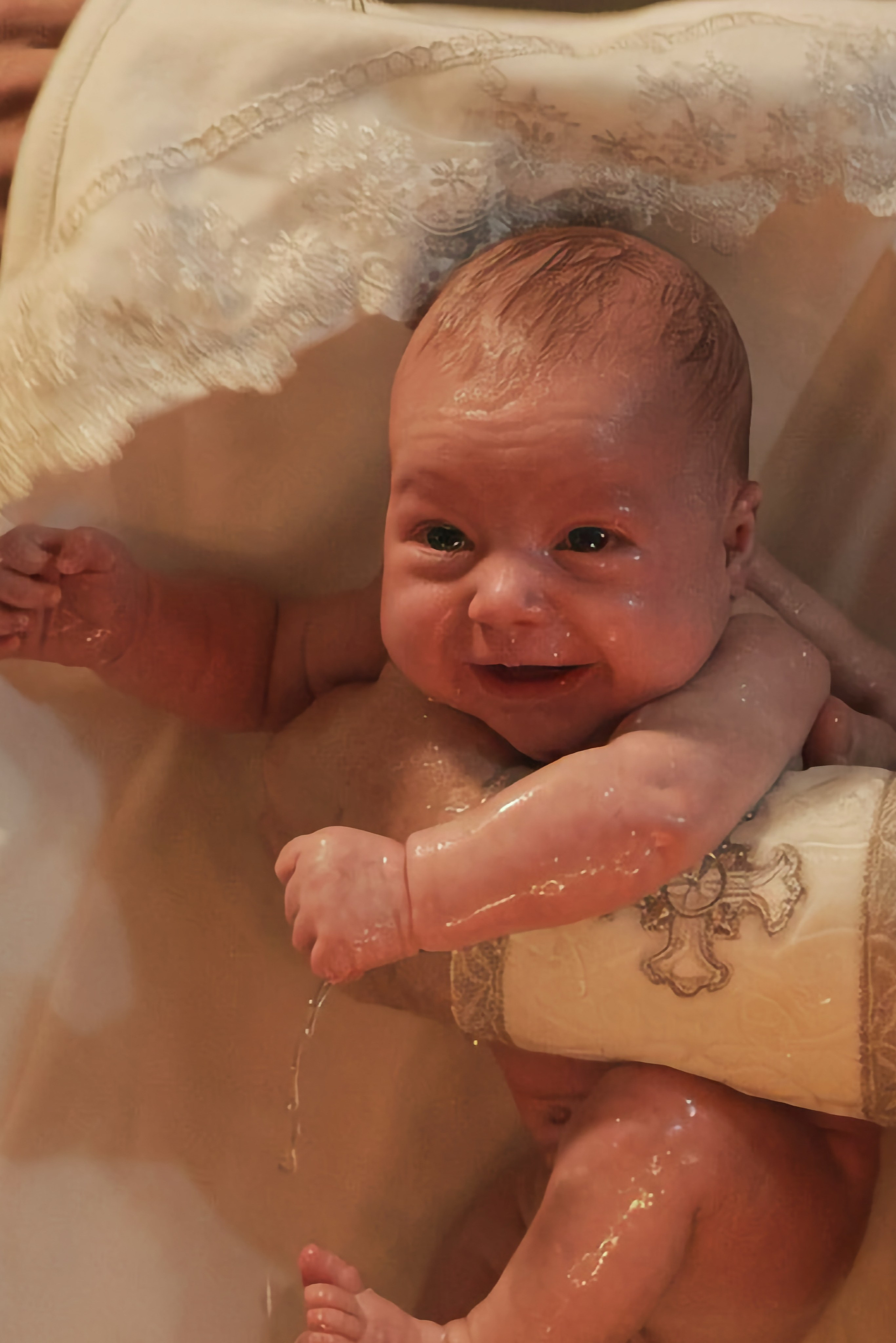 Младенец улыбается в купели сразу после погружения, вода на коже, тёплый золотистый свет — Екатеринбург, Илья Антропов