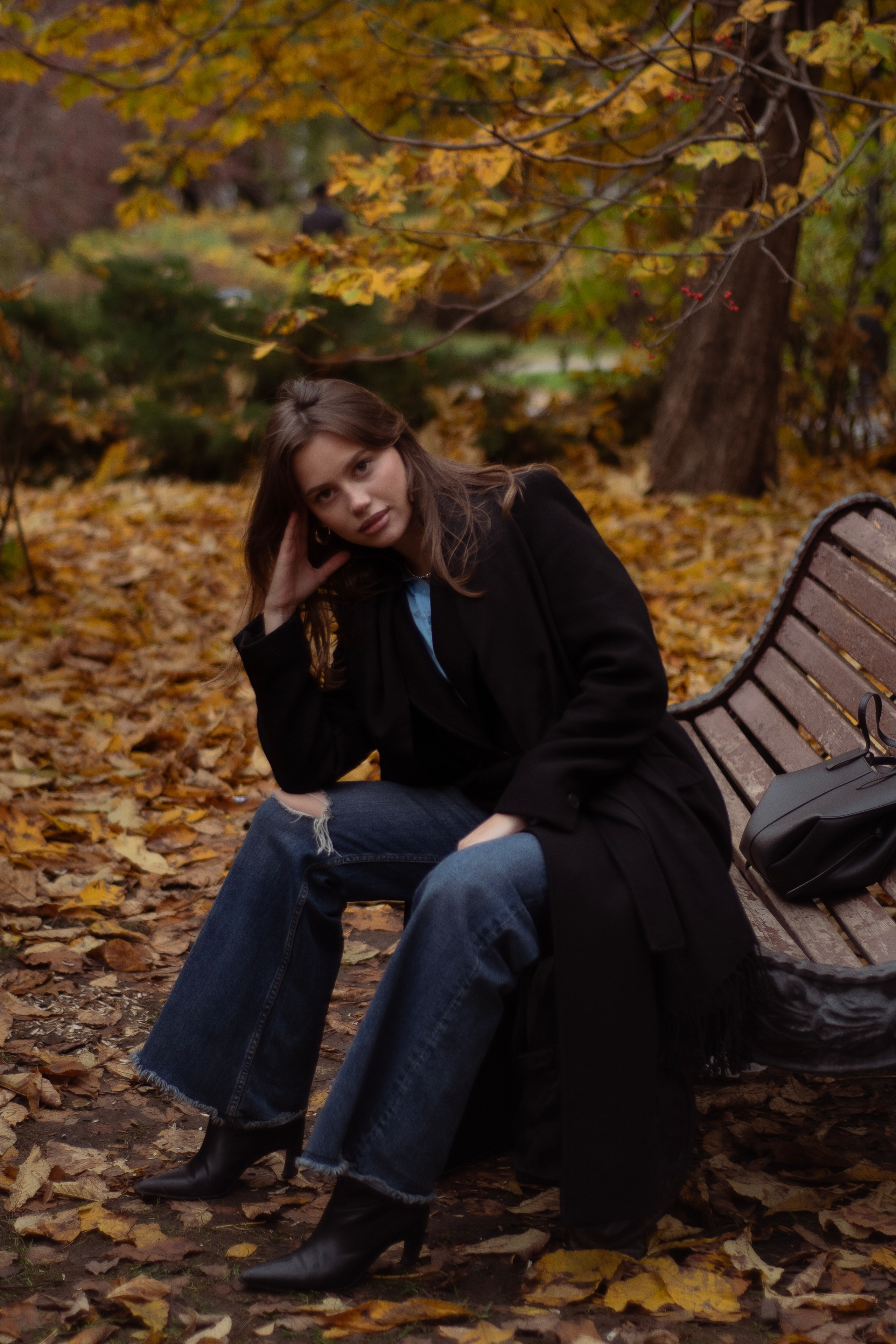 Модель в тёмном пальто, джинсах-клёш и кожаных ботинках сидит на скамейке в парке. Съёмка для бренда одежды. Стрит фотосессия . 