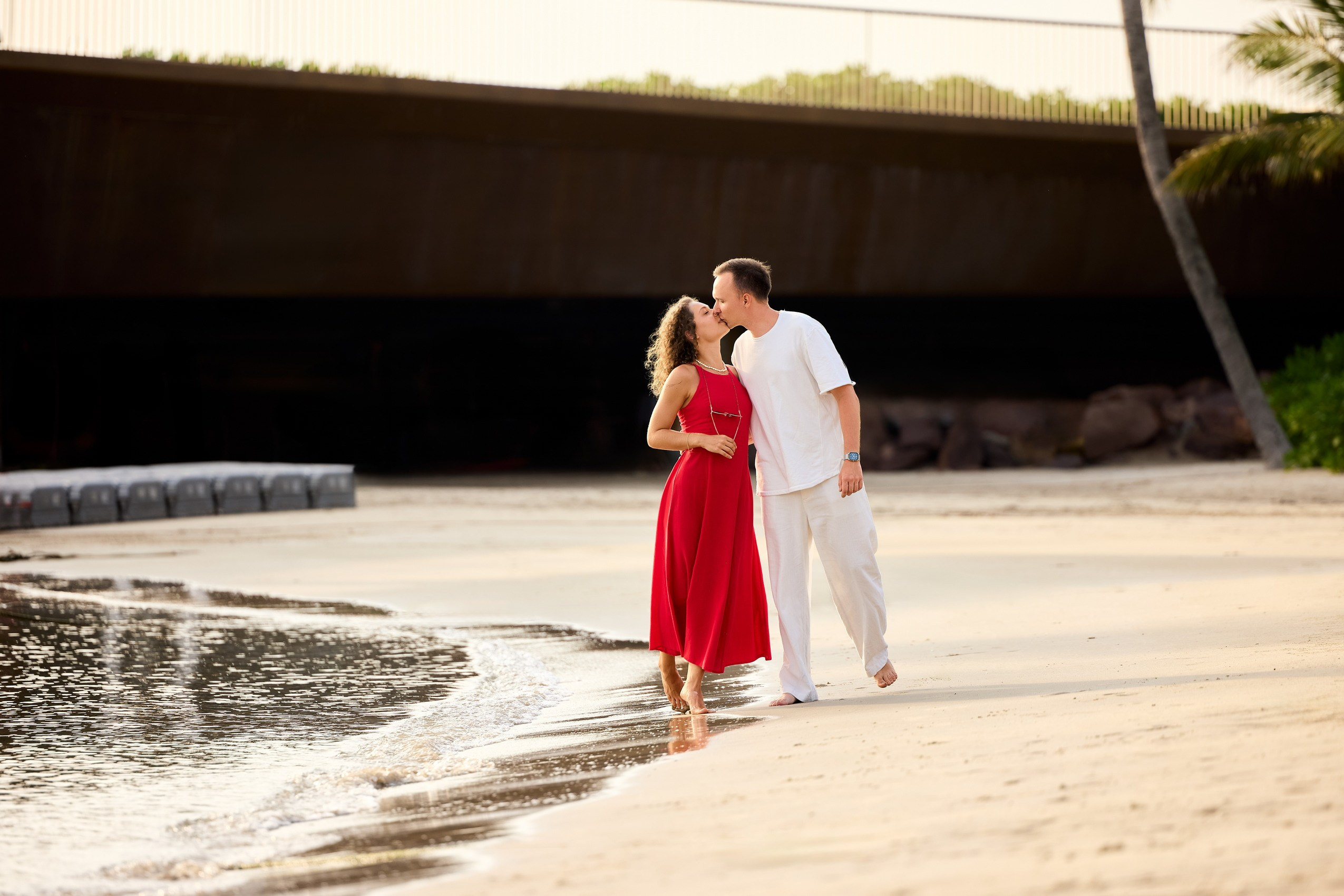 Couple sharing a kiss at the water’s edge on a sandy beach, photographed by the on‑site photographer during a couples photoshoot; tropical backdrop with palm trees and warm light, Sunset Town, Phu Quoc, Vietnam.