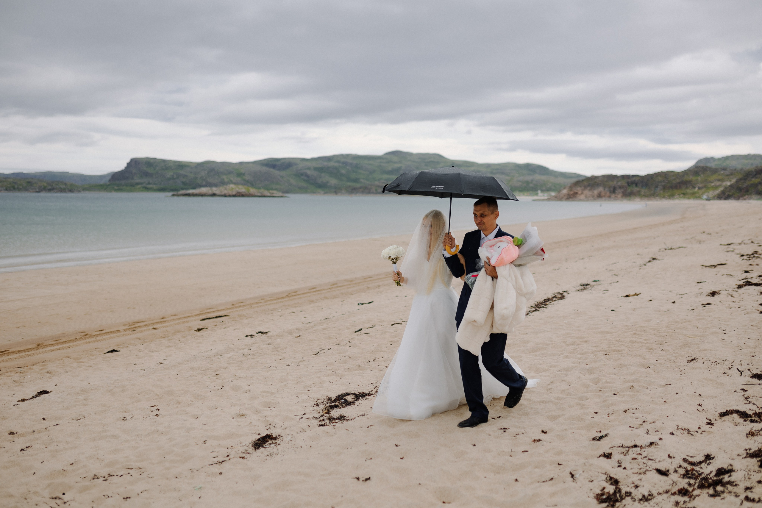 Невеста в белом платье идёт босиком по каменистому пляжу Териберки.