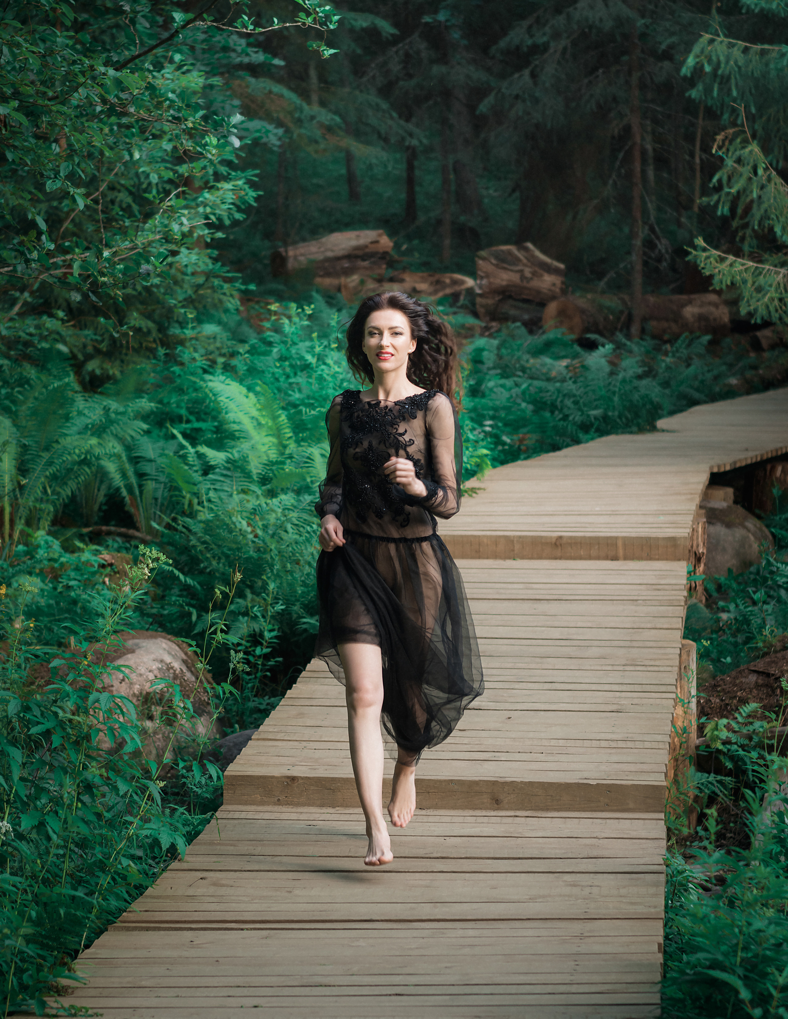 Девушка в чёрном платье бежит по деревянной дорожке в лесу