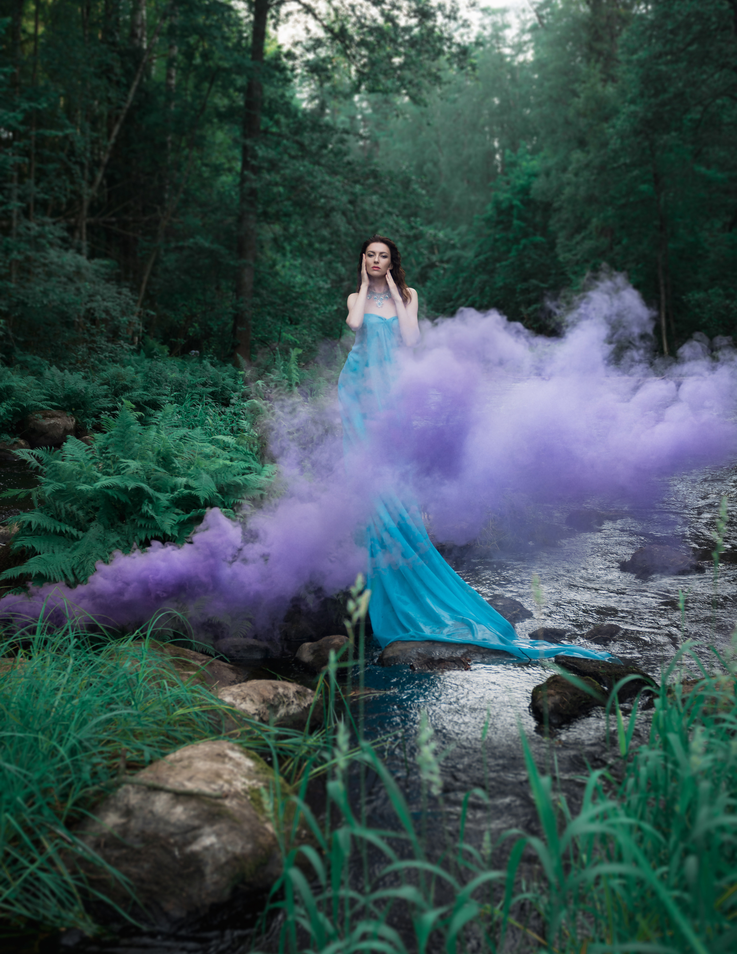 Девушка в голубом платье длинном стоит на камнях в реке, сиреневый дым