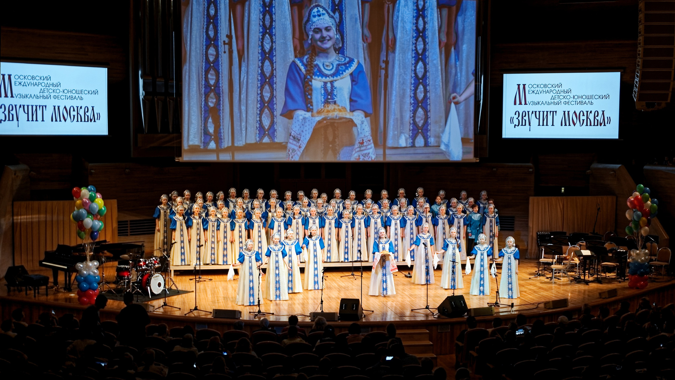 Детский хор в русских народных костюмах выступает на фестивале «Звучит Москва» - большой коллектив на сцене с экранами, съемка детского музыкального фестиваля
