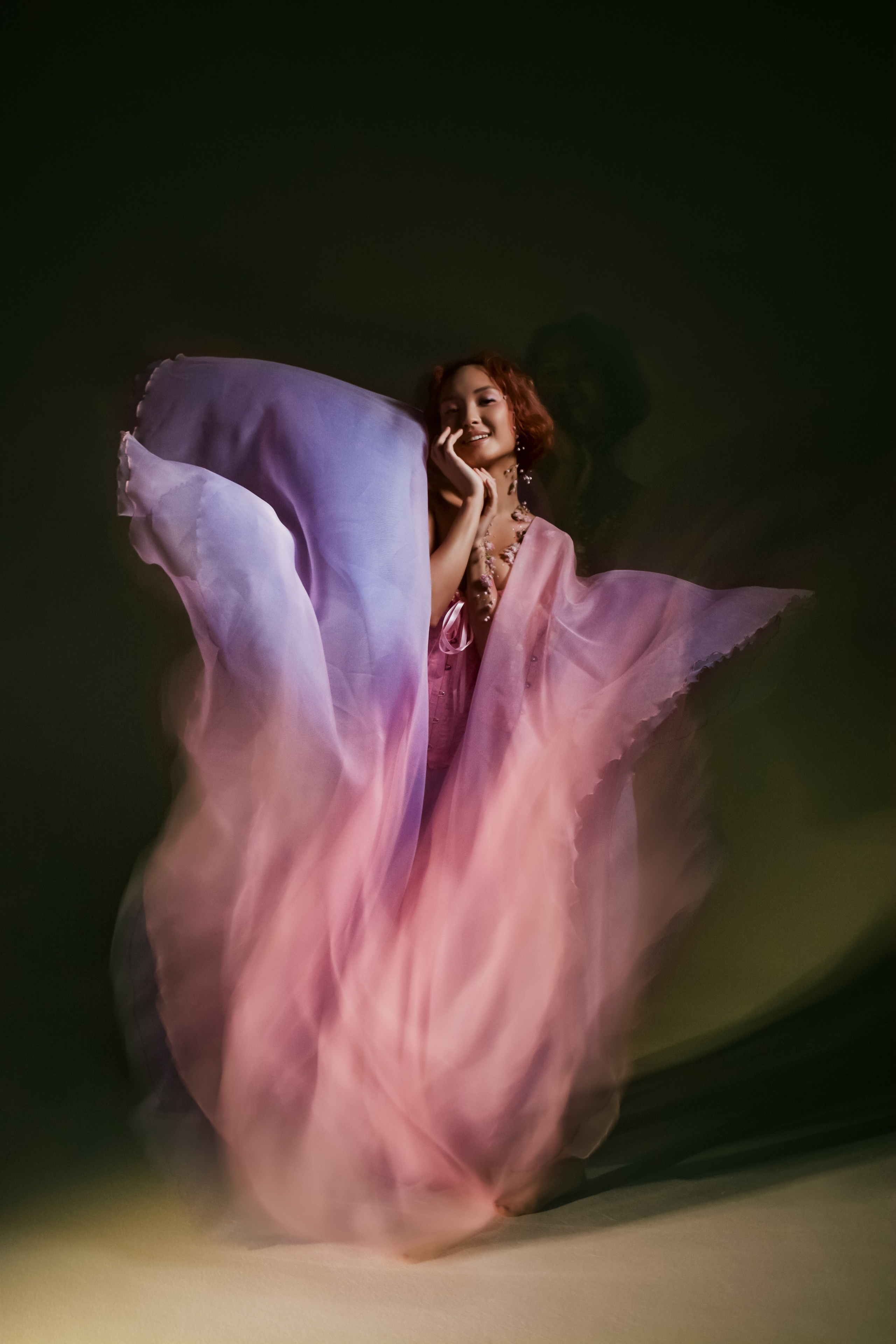 Девушка-цветок в розовом платье, олицетворяющая весеннее цветение. Ее шея и рука украшены живыми цветами, создавая гармоничный природный образ. Съемка в смешанном свете подчеркивает нежные оттенки и воздушность композиции.