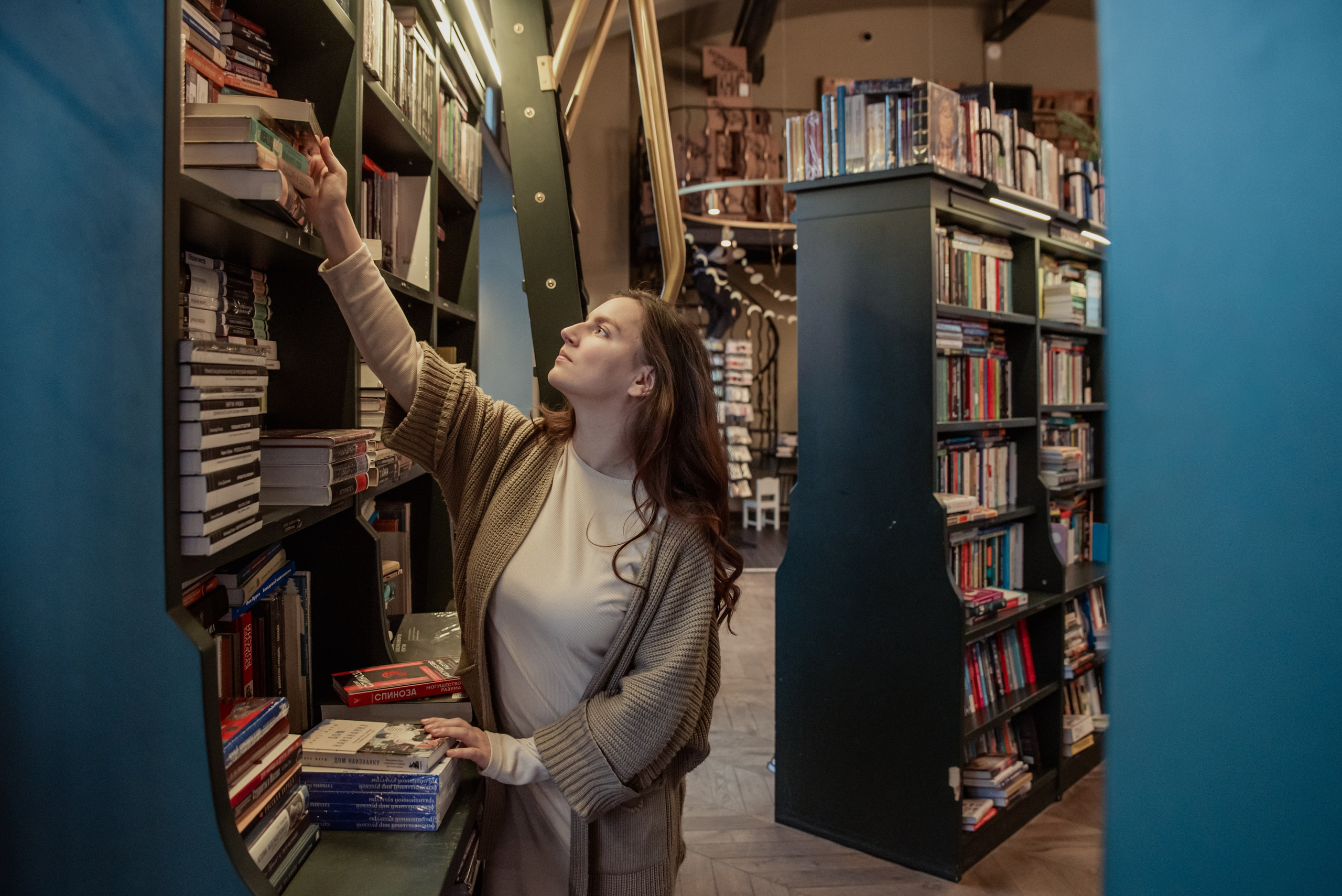 Девушка в книжном магазине напротив большого книжного шкафа тянется за книгой на полке.