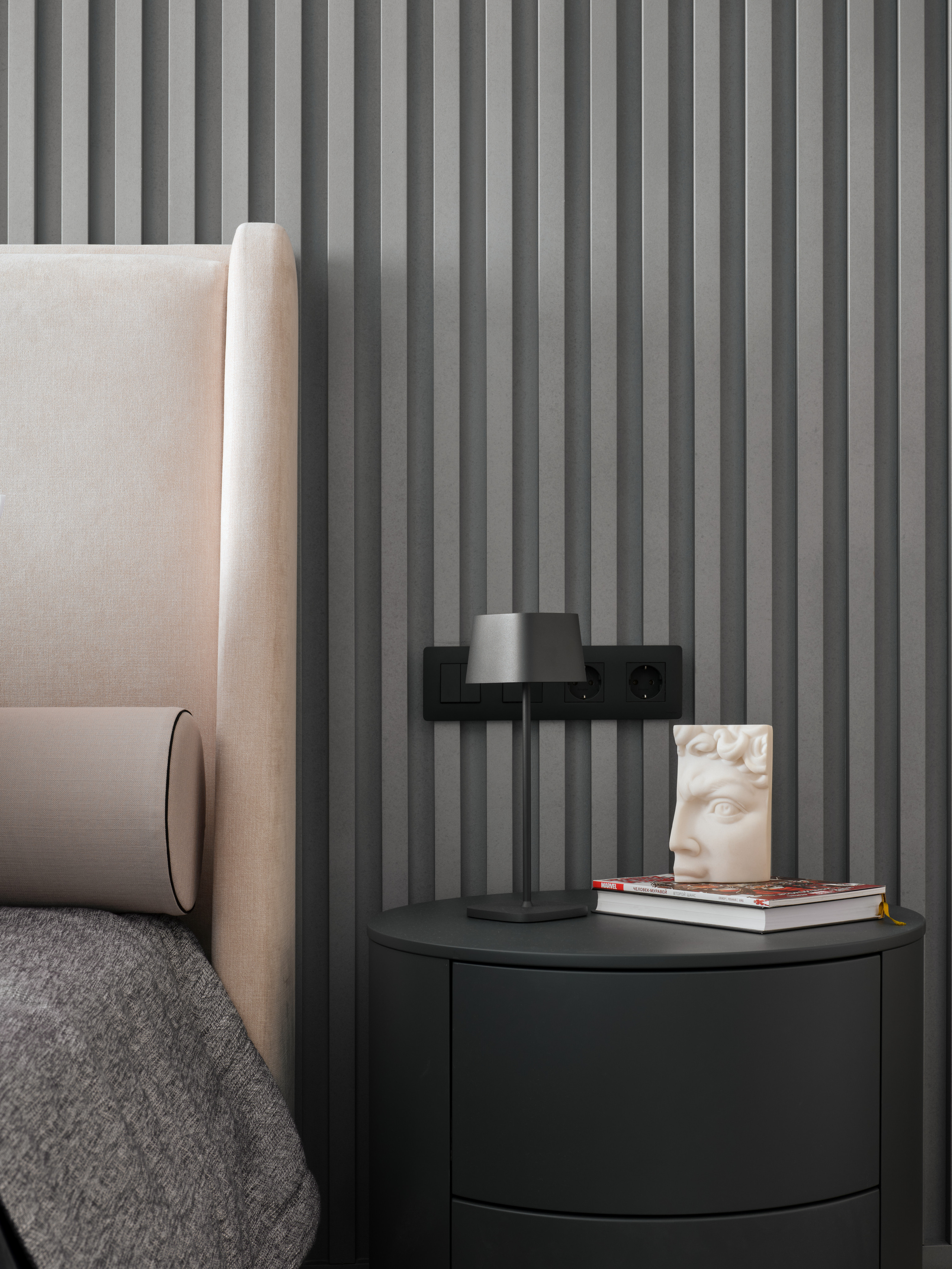 Спальня с мягким изголовьем кровати, серой акцентной стеной с вертикальными панелями и прикроватной тумбой, интерьер Екатерины Мищенко, фотограф Сергей Денисенко, современный стиль с акцентом на текстуры и детали.