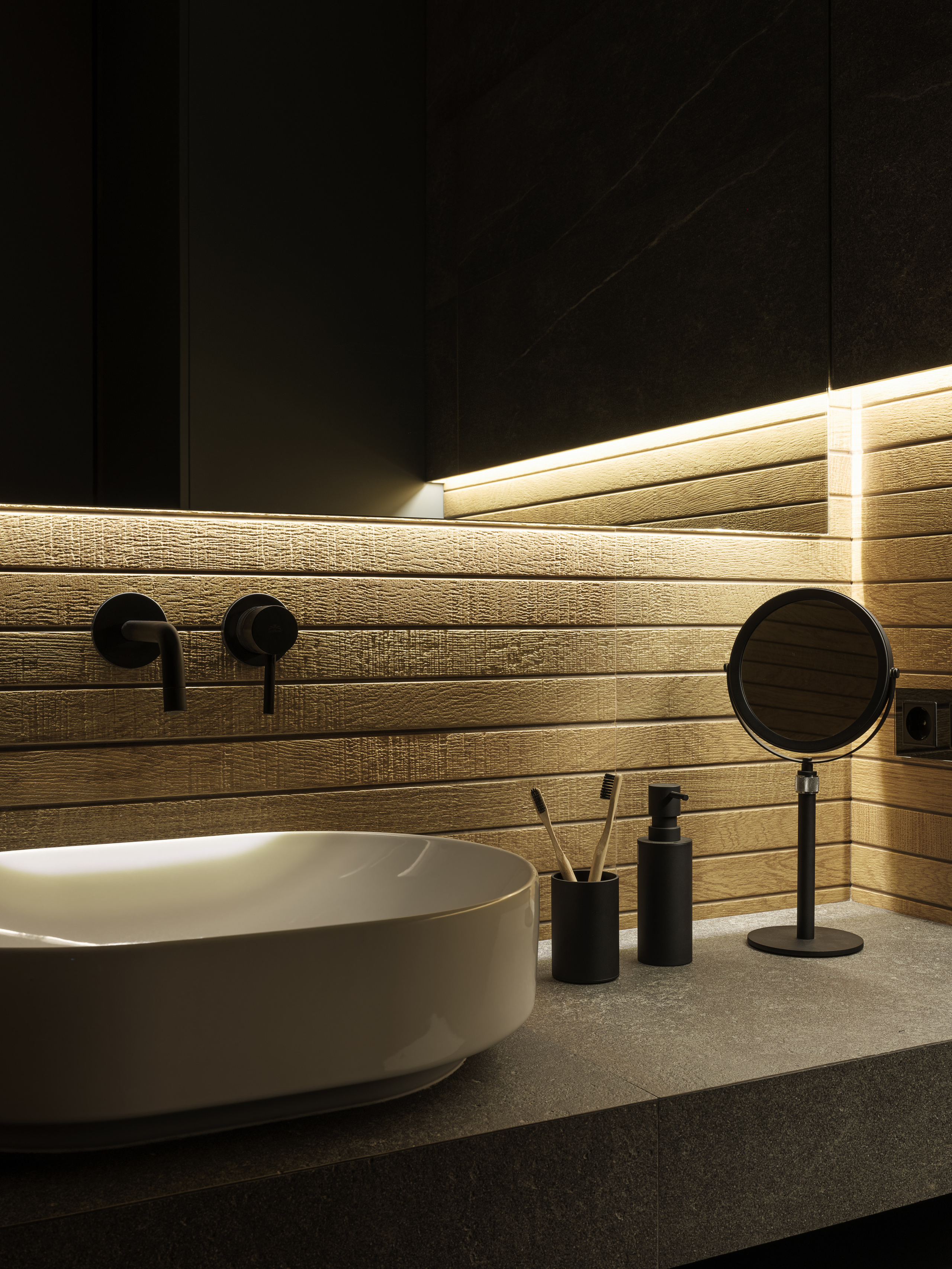 Фрагмент ванной комнаты с раковиной на каменной столешнице, деревянными панелями, подсветкой под зеркалом, аксессуарами в черном минимализме и круглым косметическим зеркалом, дизайн Алёна Сверлова, съемка Сергей Денисенко.