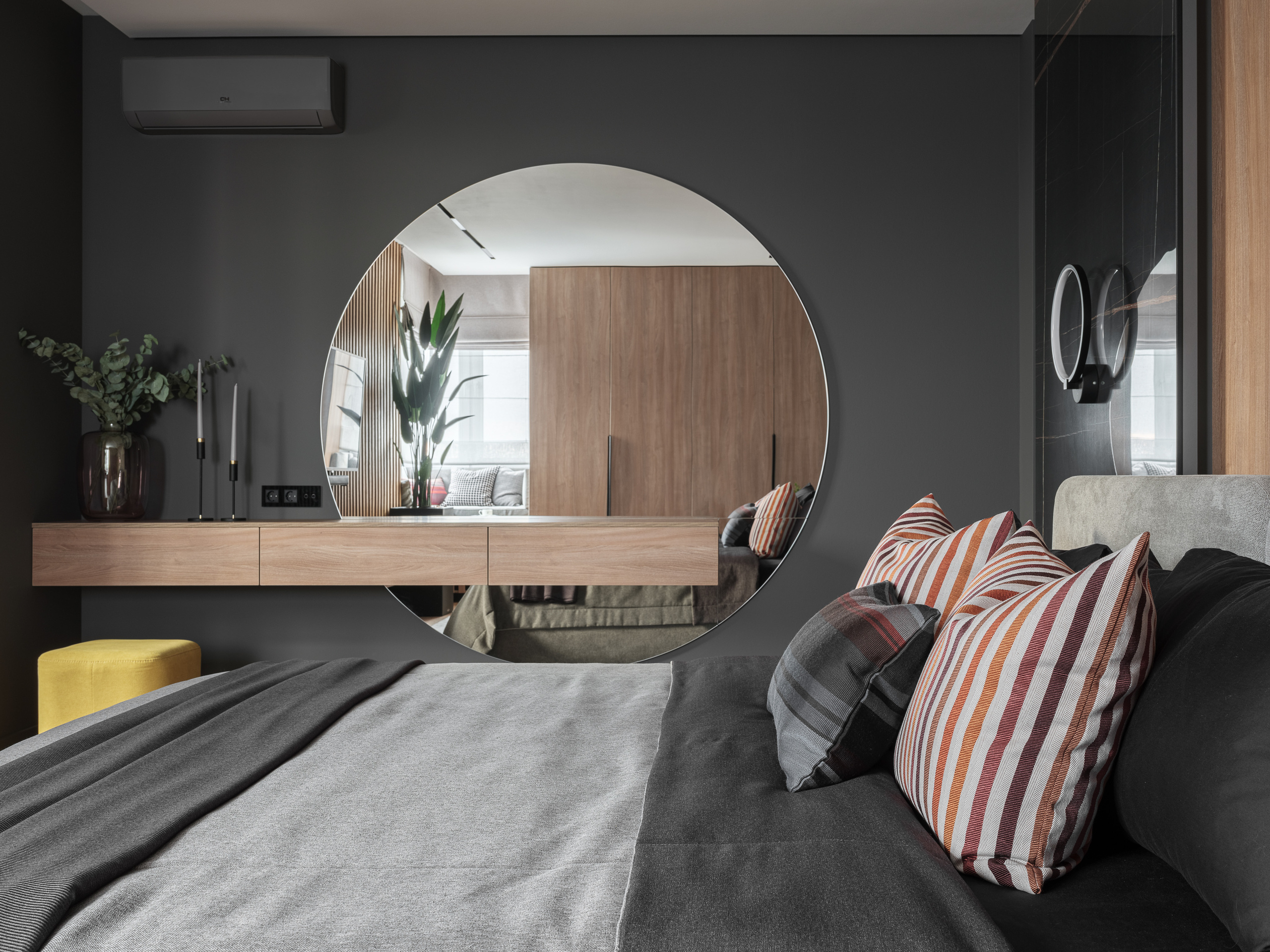 Дизайнерская спальня в серых и деревянных тонах с круглым зеркалом на стене, серым текстилем на кровати и полосатыми подушками. Дизайнер Алёна Сверлова, фотограф Сергей Денисенко.