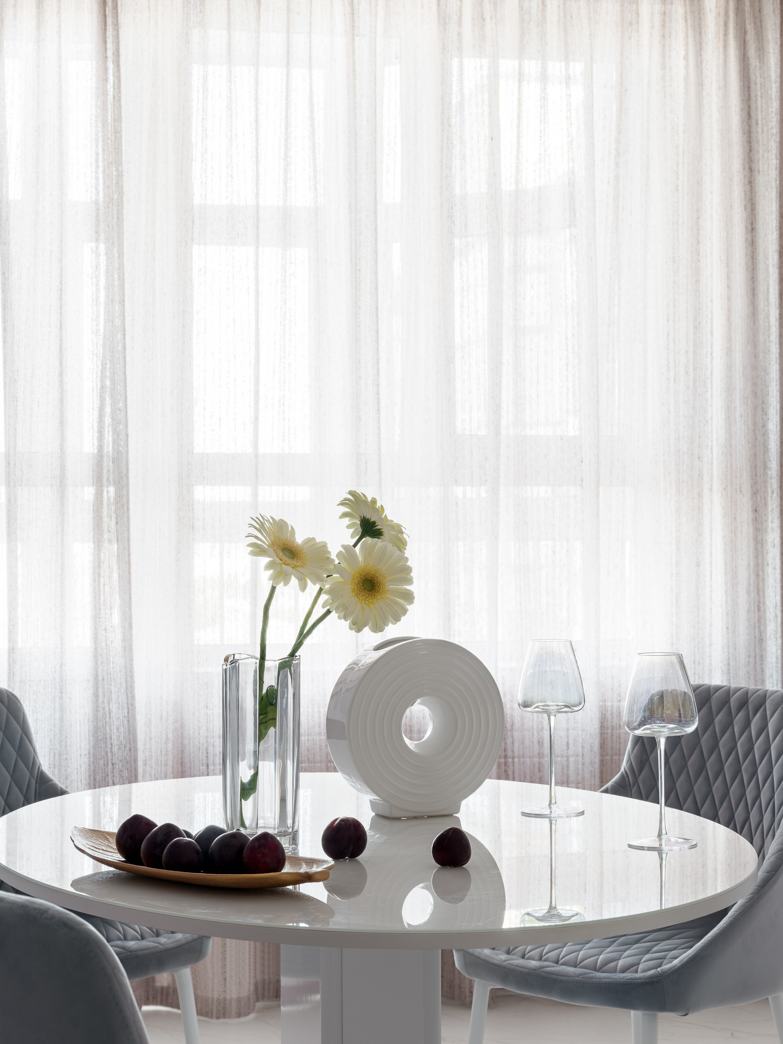 Светлая столовая с большими окнами в легких полупрозрачных шторах, белый круглый стол с цветами и фруктами, мягкие стулья серого цвета; интерьер создан дизайнером Натальей Бунеевой, стилист Наталья Якушева, фотограф Сергей Денисенко.