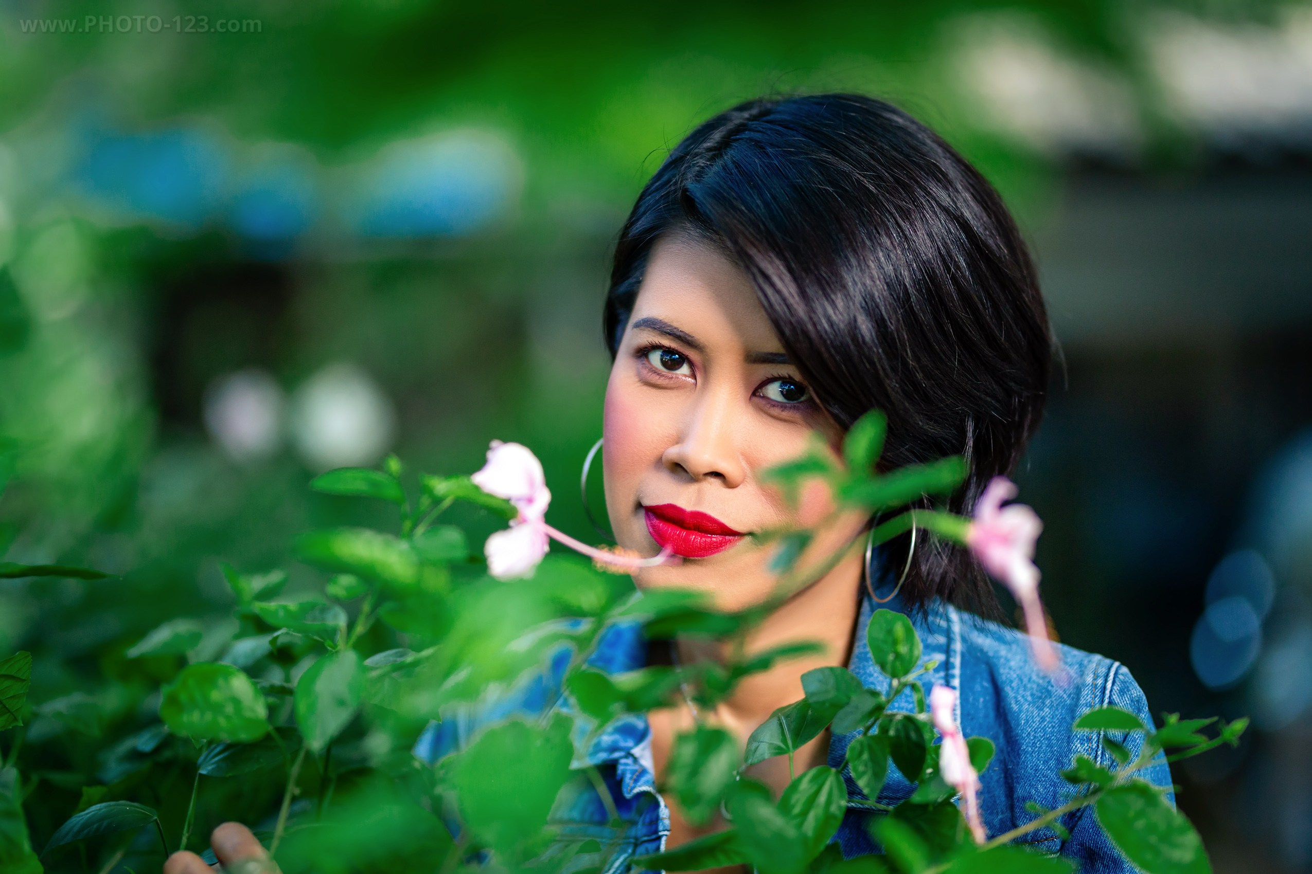 Outdoor portrait of a woman with short dark hair and red lipstick looking through green leaves with small pink flowers, wearing a denim jacket and hoop earrings, shallow depth of field, soft natural light, lifestyle portrait photography in a garden setting