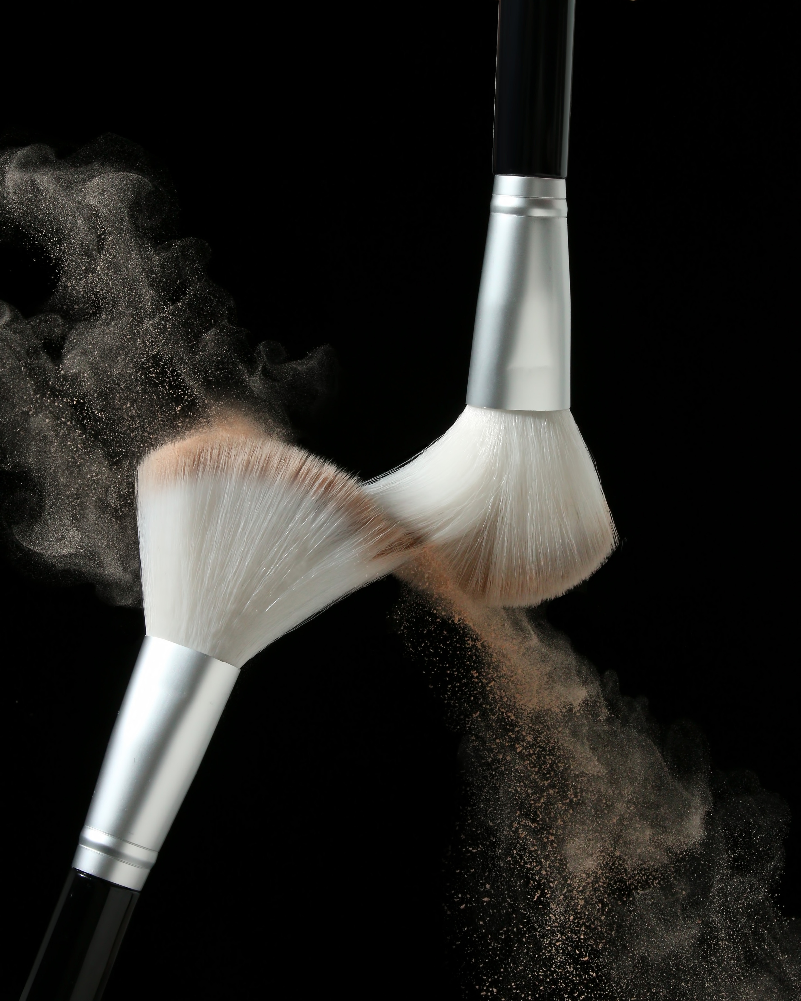 Две кисти для пудры на черном фоне, между ними - летящая пудра