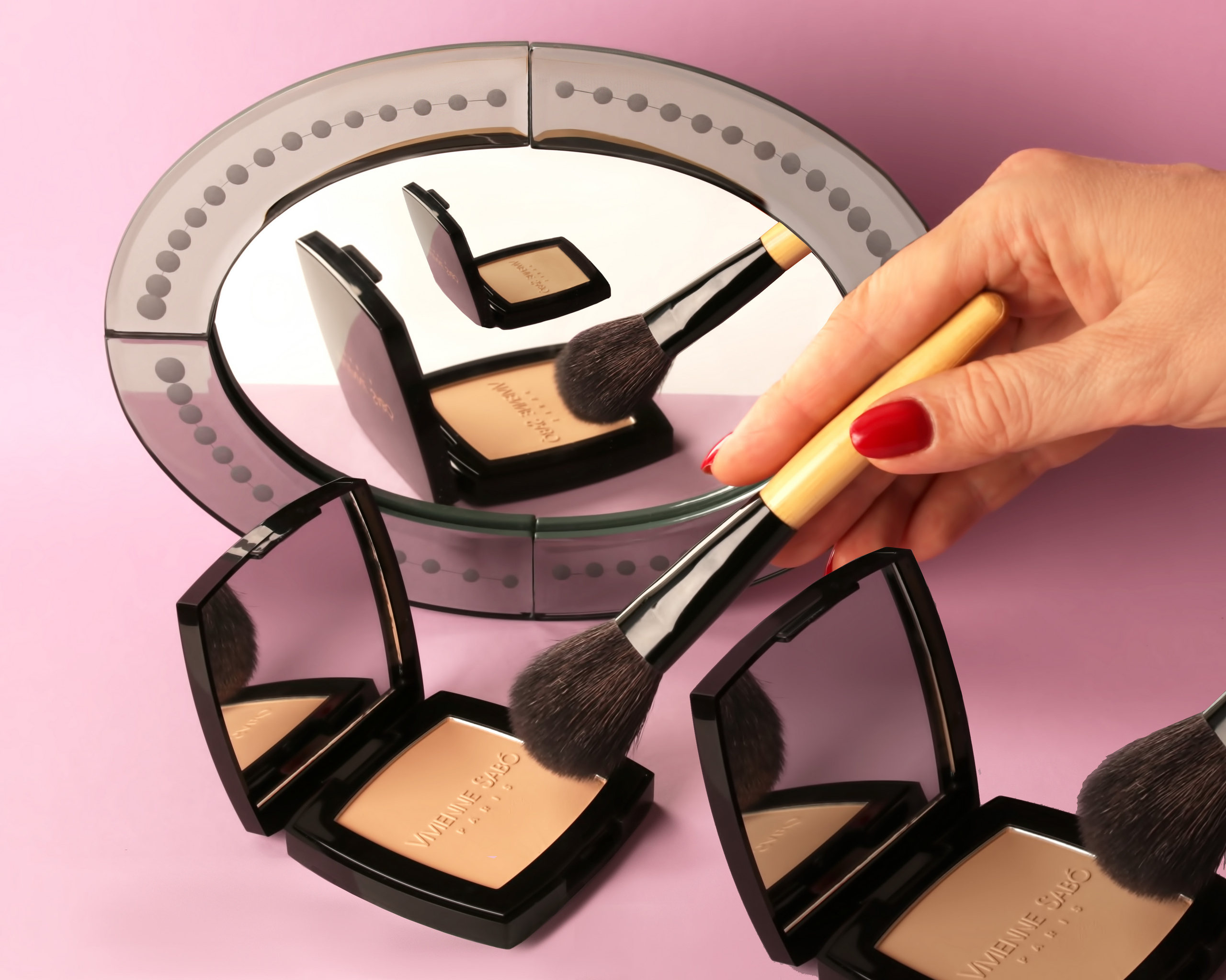 Пудреница Vivienne Sabo отражается в зеркале, над ней - рука с кисточкой