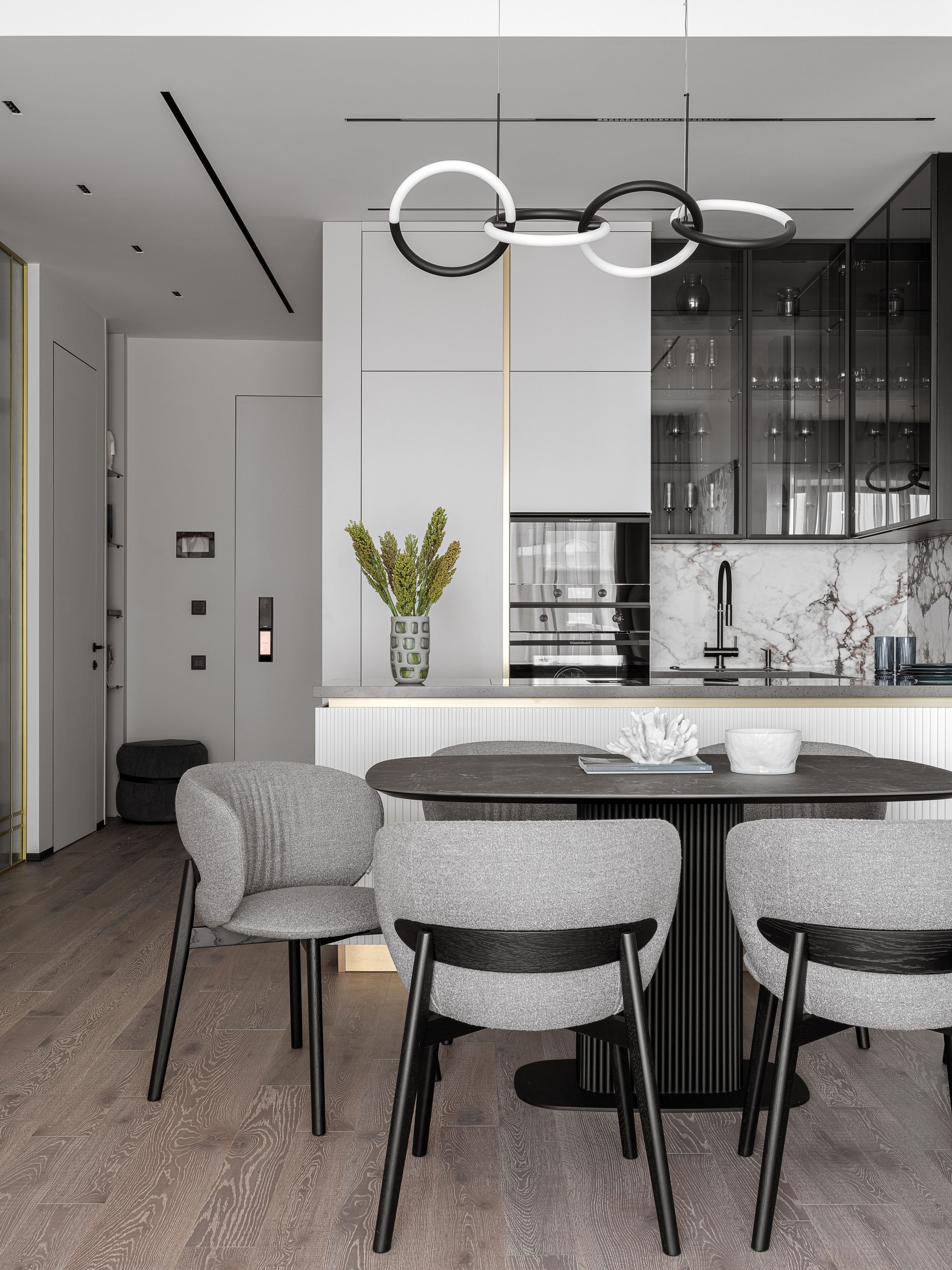 Современная кухня-гостиная в проекте Сочи: обеденный стол с мягкими серыми стульями, подвесной светильник с кольцами, белые фасады кухни и акцентная мраморная отделка. Дизайн: Ренат Байчуков, Алеся Кириенко; стилист Екатерина Ракитина; фото Сергея Денисенко.
