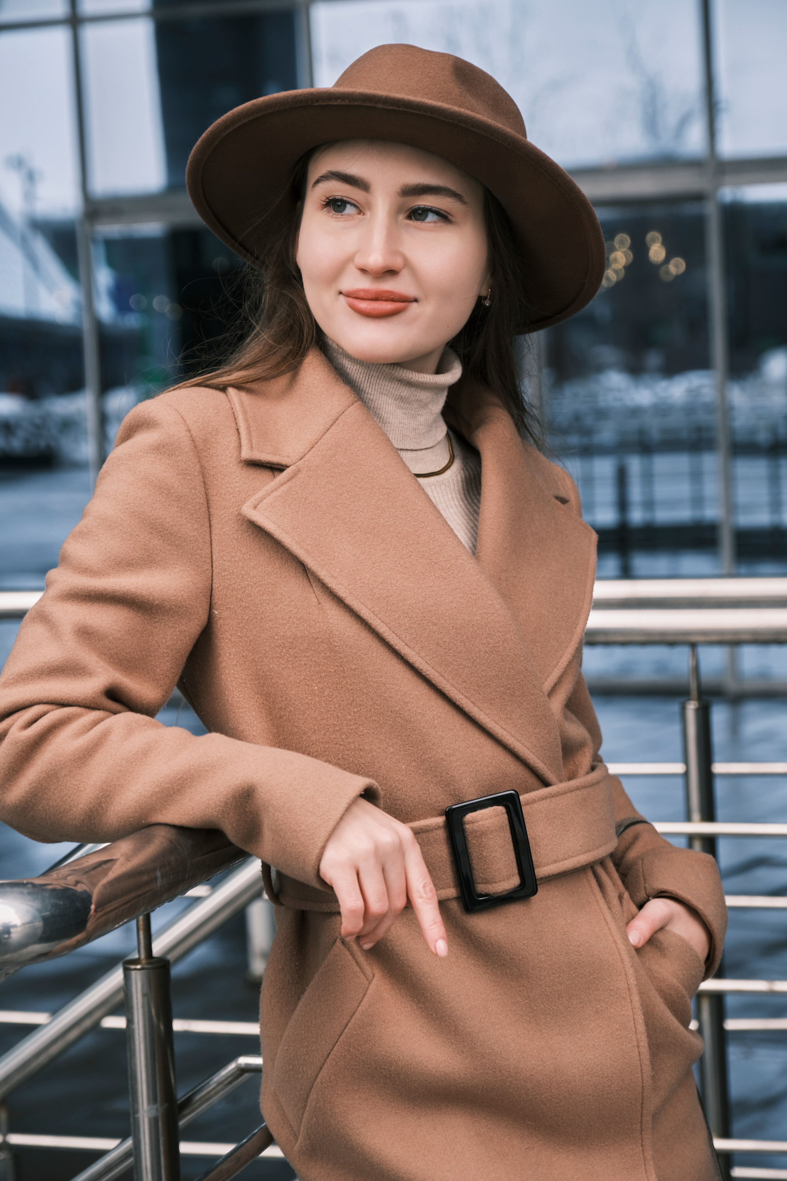 Женская фотосессия в Москва-Сити: Девушка в коричневом тренче и шляпе стоит у металлических перил на фоне современной городской архитектуры.