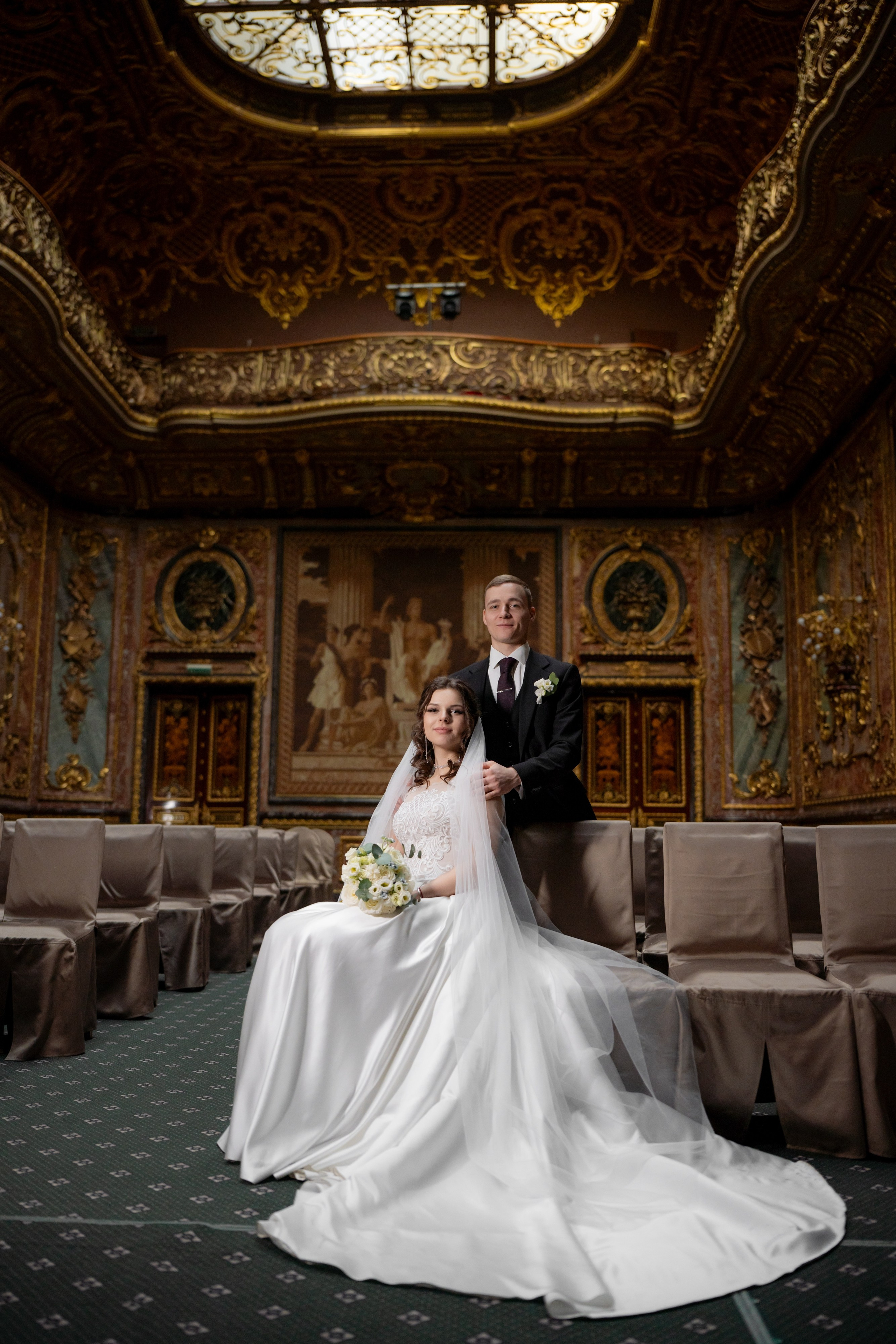 Свадебная фотосессия в Санкт-Петербурге в Особняке Половцева или Дом Архитектора. Невеста сидит на стуле, а жених стоит рядом за стулом в зале театра.