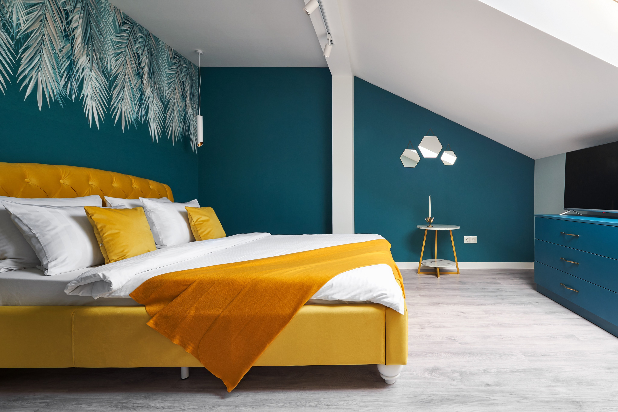 Желтая кровать с белым постельным бельем в спальне-мансарде с бирюзовыми стенами.