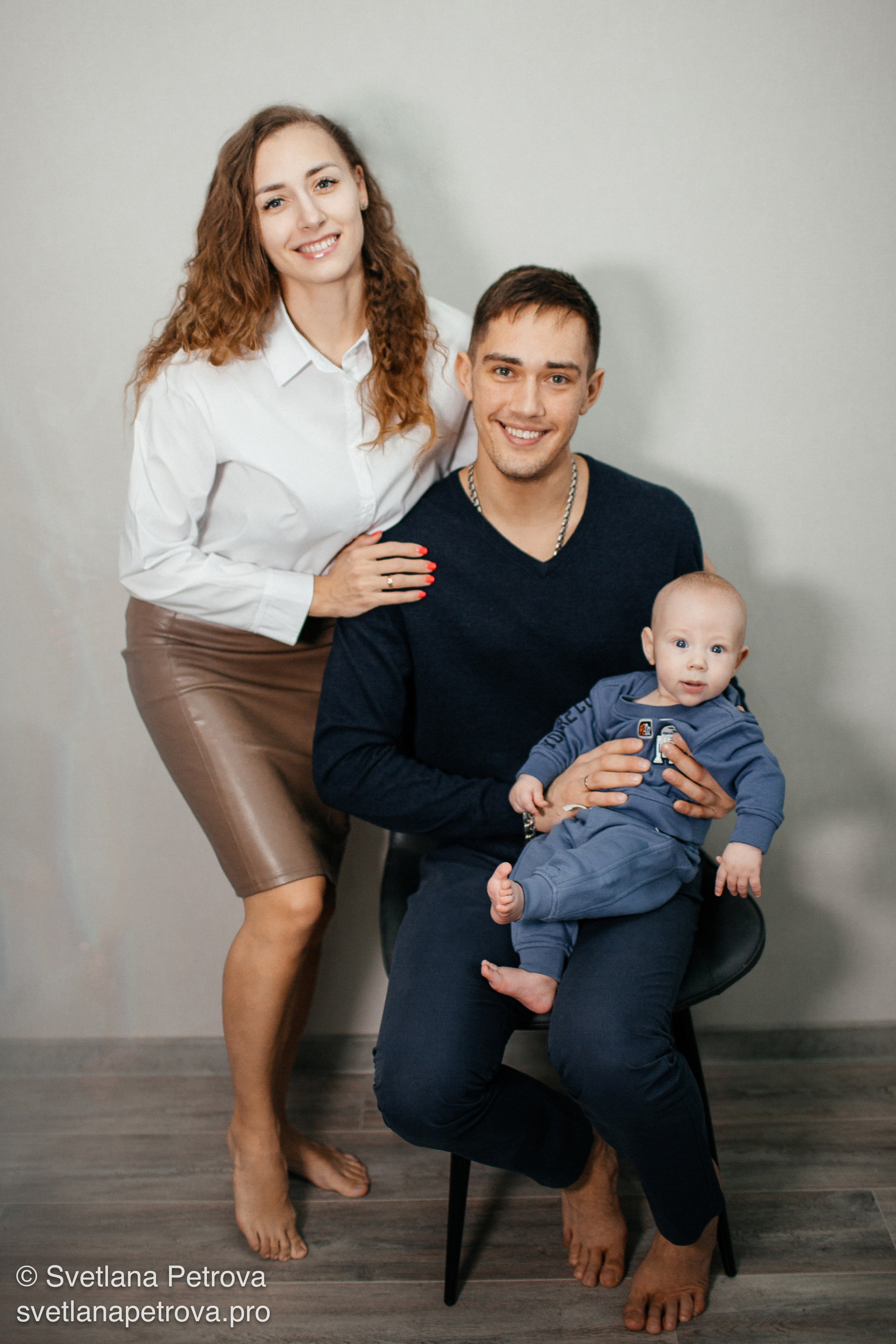 Родители с младенцем — светлый семейный портрет. Мама обнимает папу, малыш на руках — радость в кадре. Семья втроём — момент нежности и счастья. Младенец в объятиях папы, мама рядом улыбается. Тёплый семейный снимок: родители и их малыш. Улыбки мамы и папы, малыш изучает камеру. Семейный портрет: родители и кроха в уютной студии.