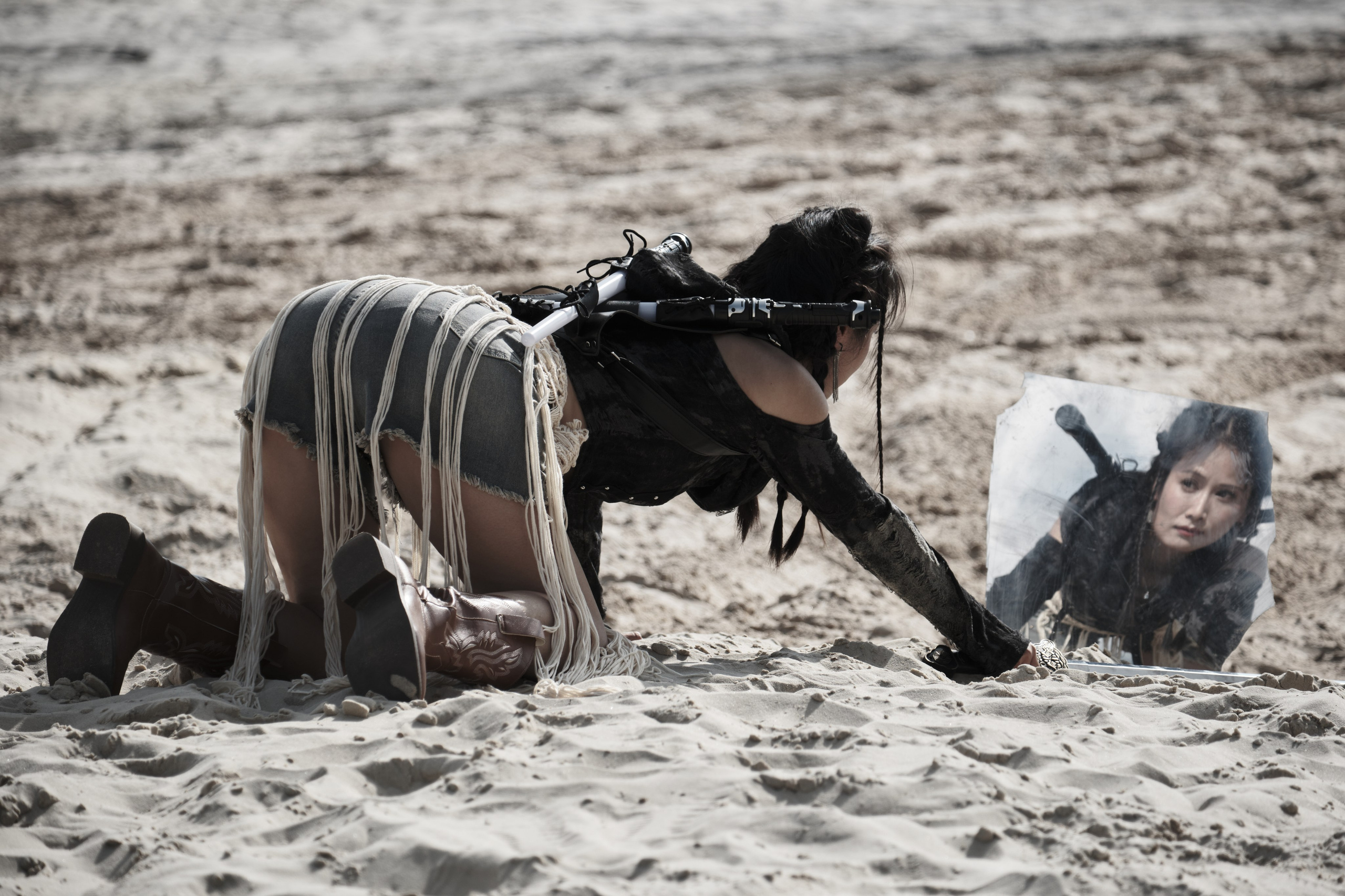 Девушка азиатской внешности ползет по песку и смотрит в зеркало в стиле пост апокалипсис