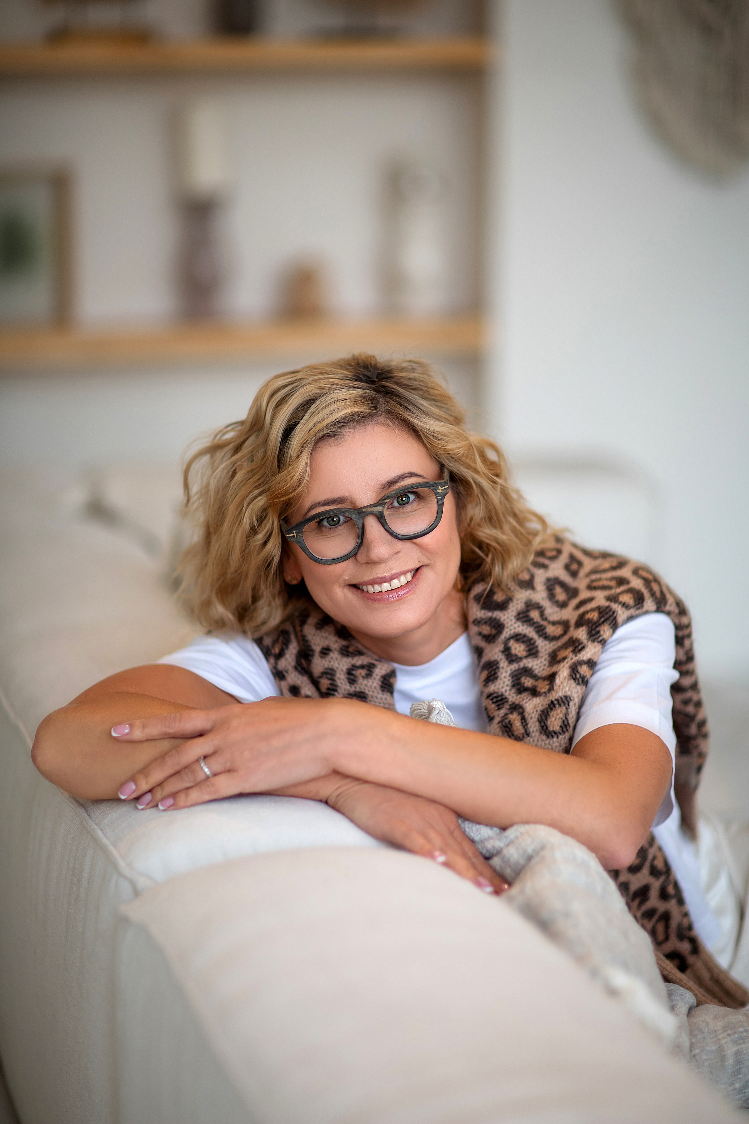 женщина в очках в леопардовом свитере сидит в светлой комнате на бежевом диване