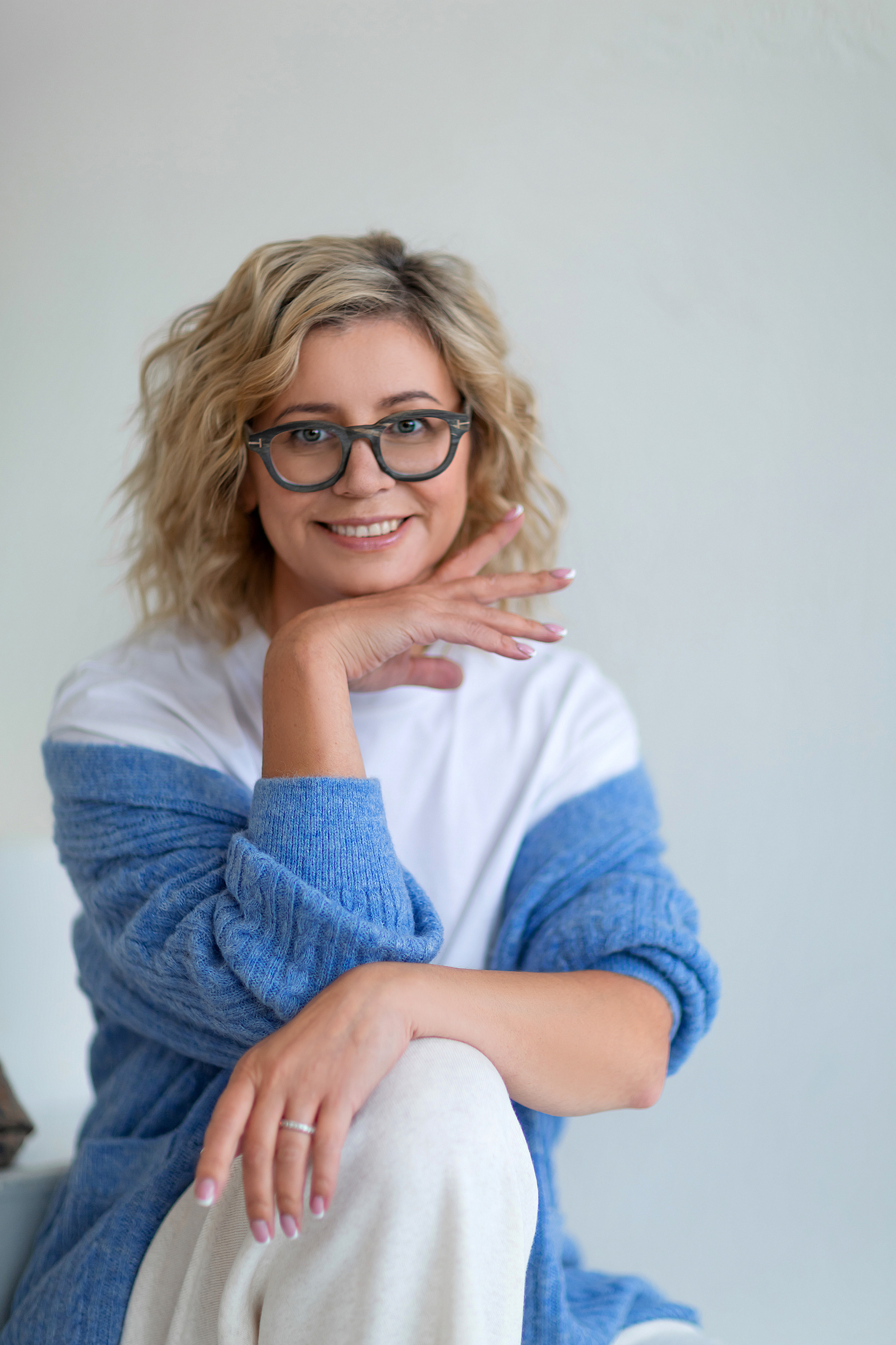 женщина с каре в голубом свитере сидит на ступеньках на белом фоне