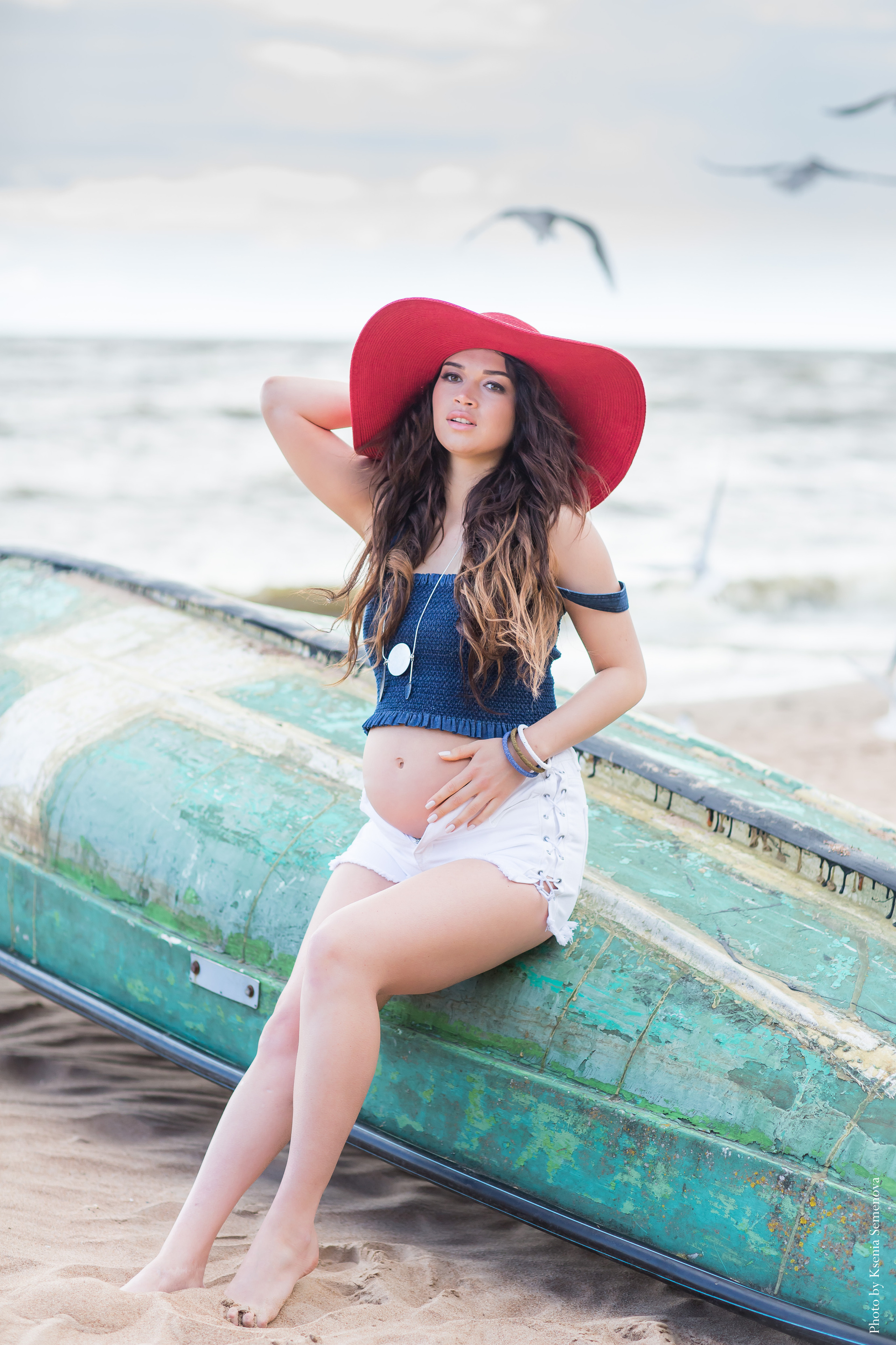 беременная девушка в синем топе, белых шортах и красной шляпе сидит на перевернутой лодке зелёного цвета на побережье финского залива