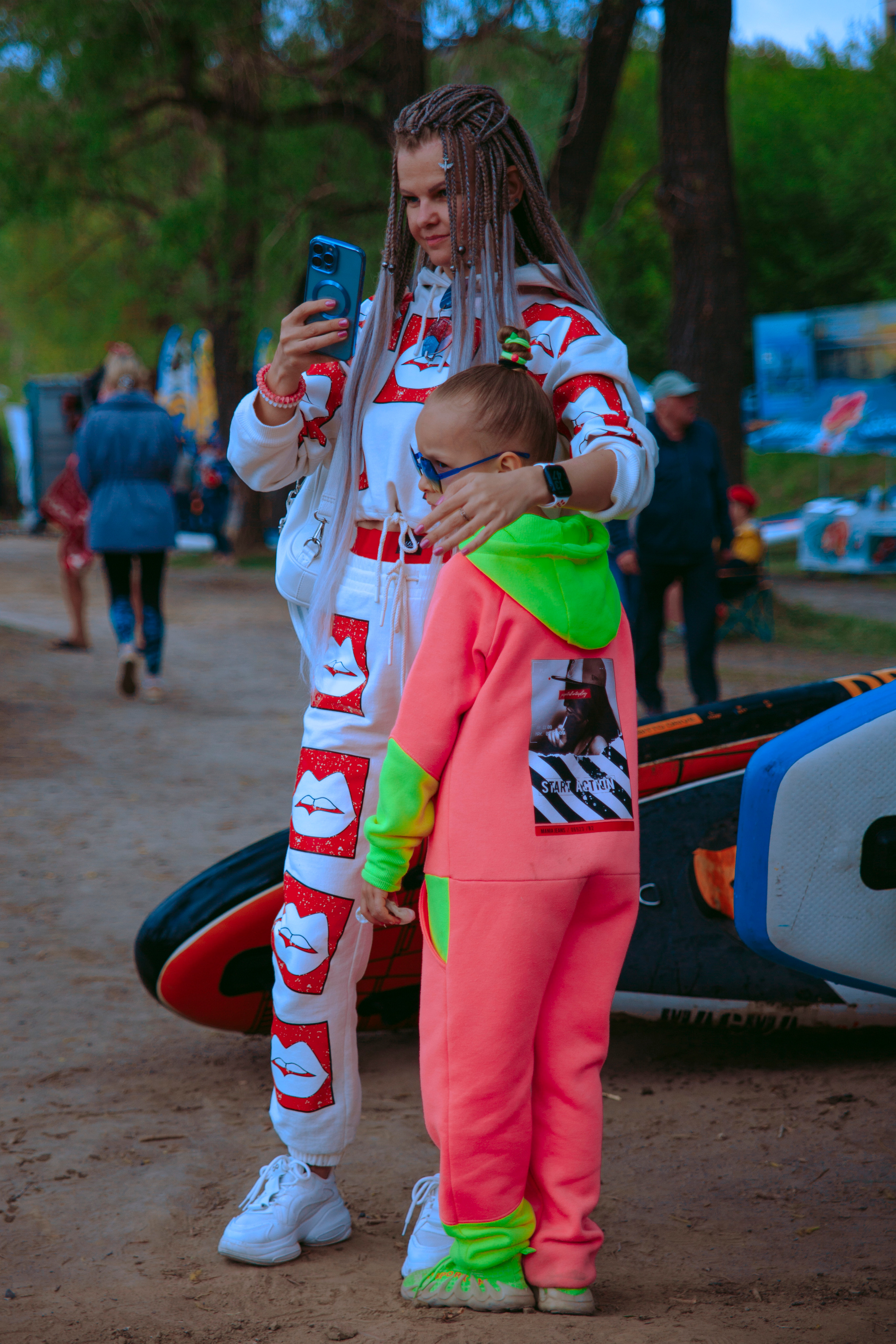 Яркая девушка с афро-косами в белом спортивном костюме и с дочкой в розовом костюме снимает что-то на телефон.