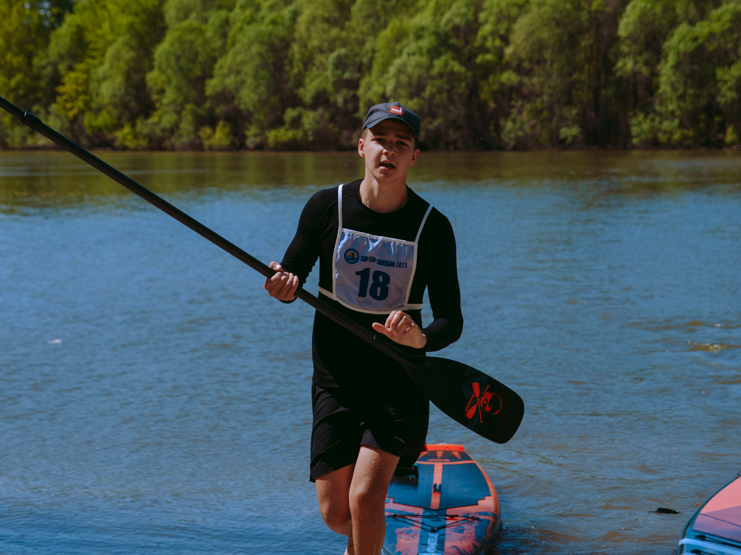 Парень в гидрокостюме с веслом идет по берегу.