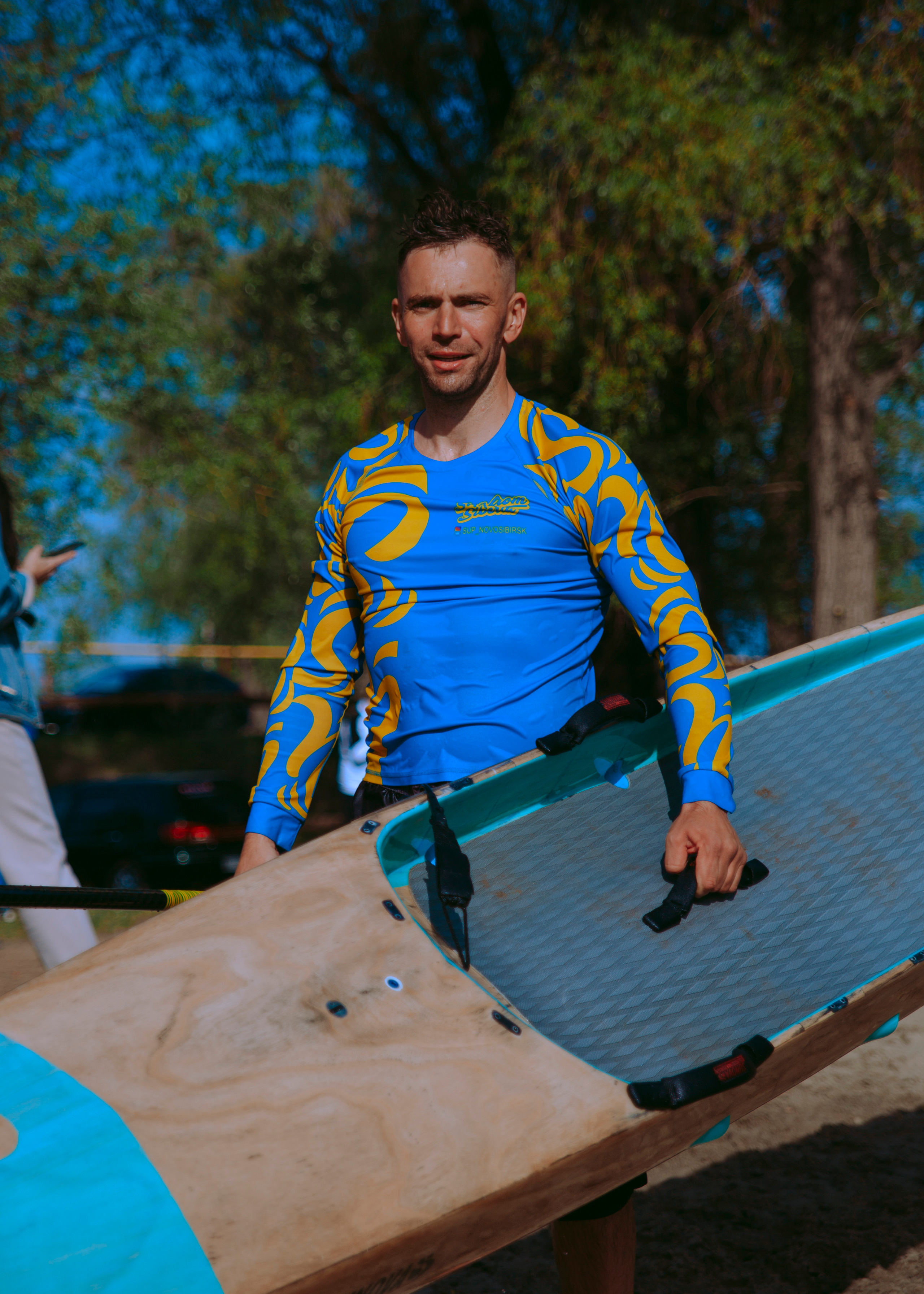 Мужчина серфер в рашгарде в гидрокостюме стоит на берегу.