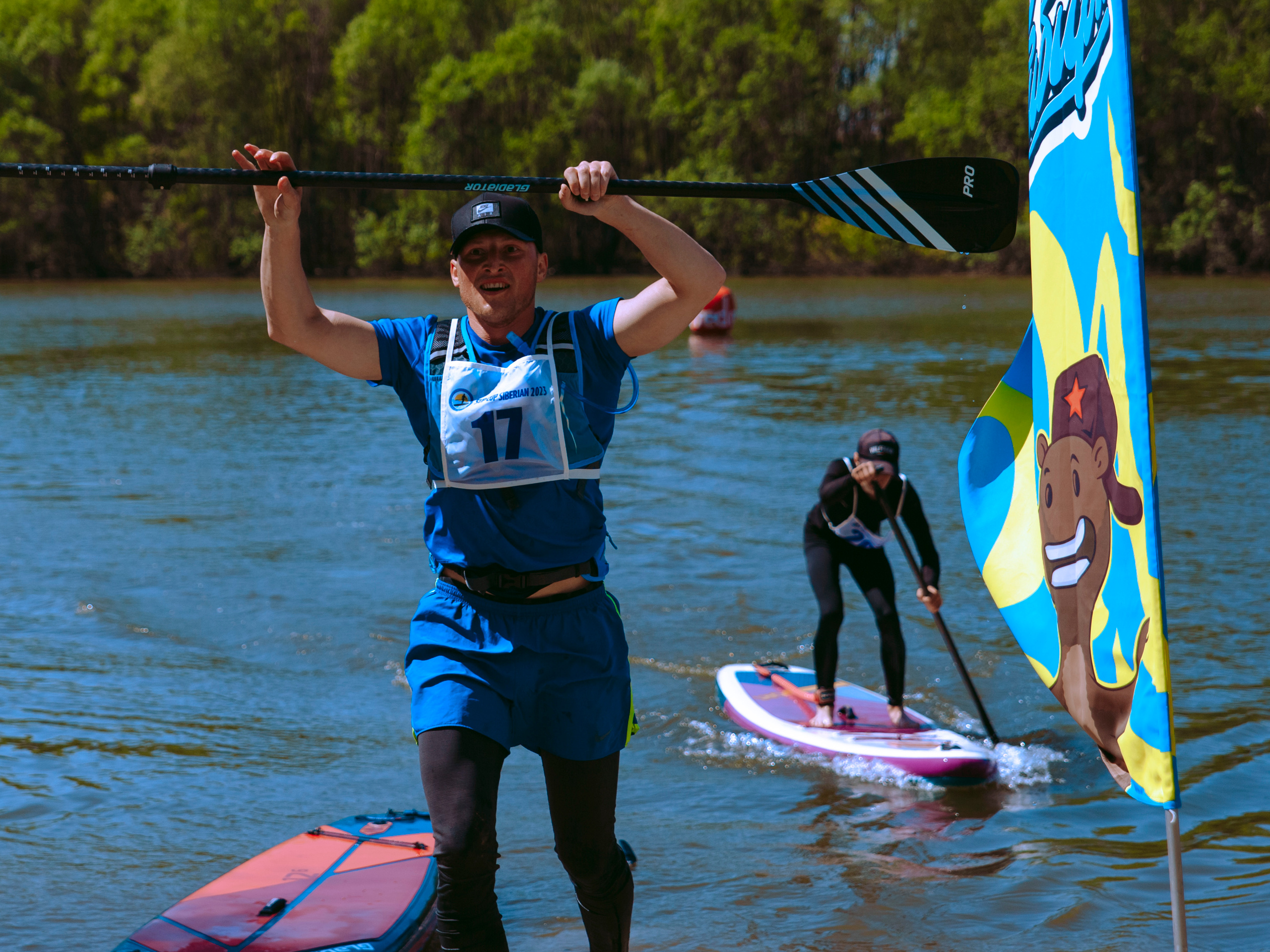 Мужчина поднимает весло и радуется победе на фоне мужчины, плывущего на сапе.