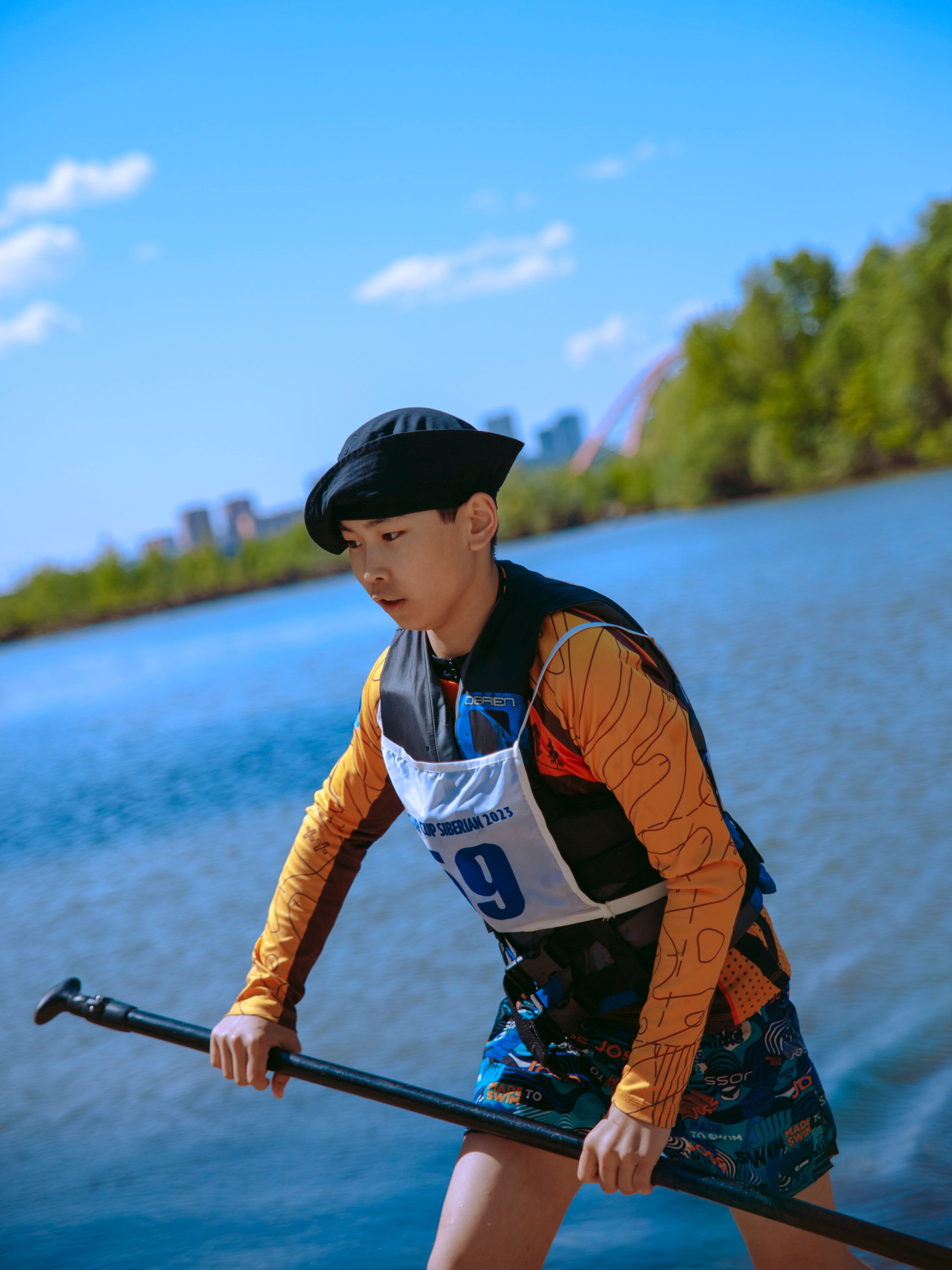 Азиатский парень в панаме с подвернутыми краями идет с веслом в руках на фоне реки.