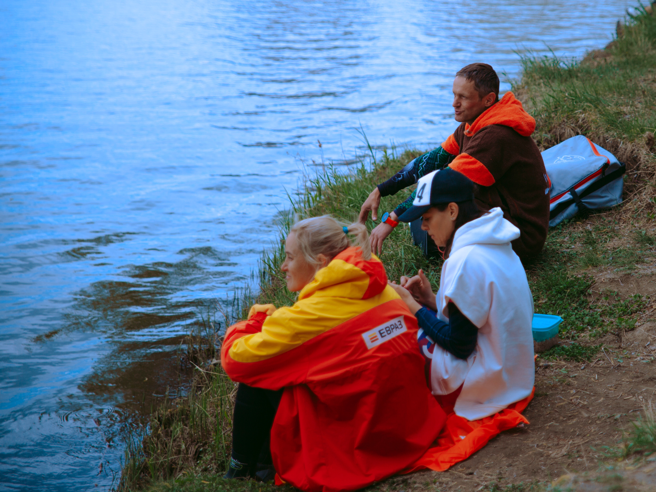 Люди в куртках ветровках сидят на берегу.