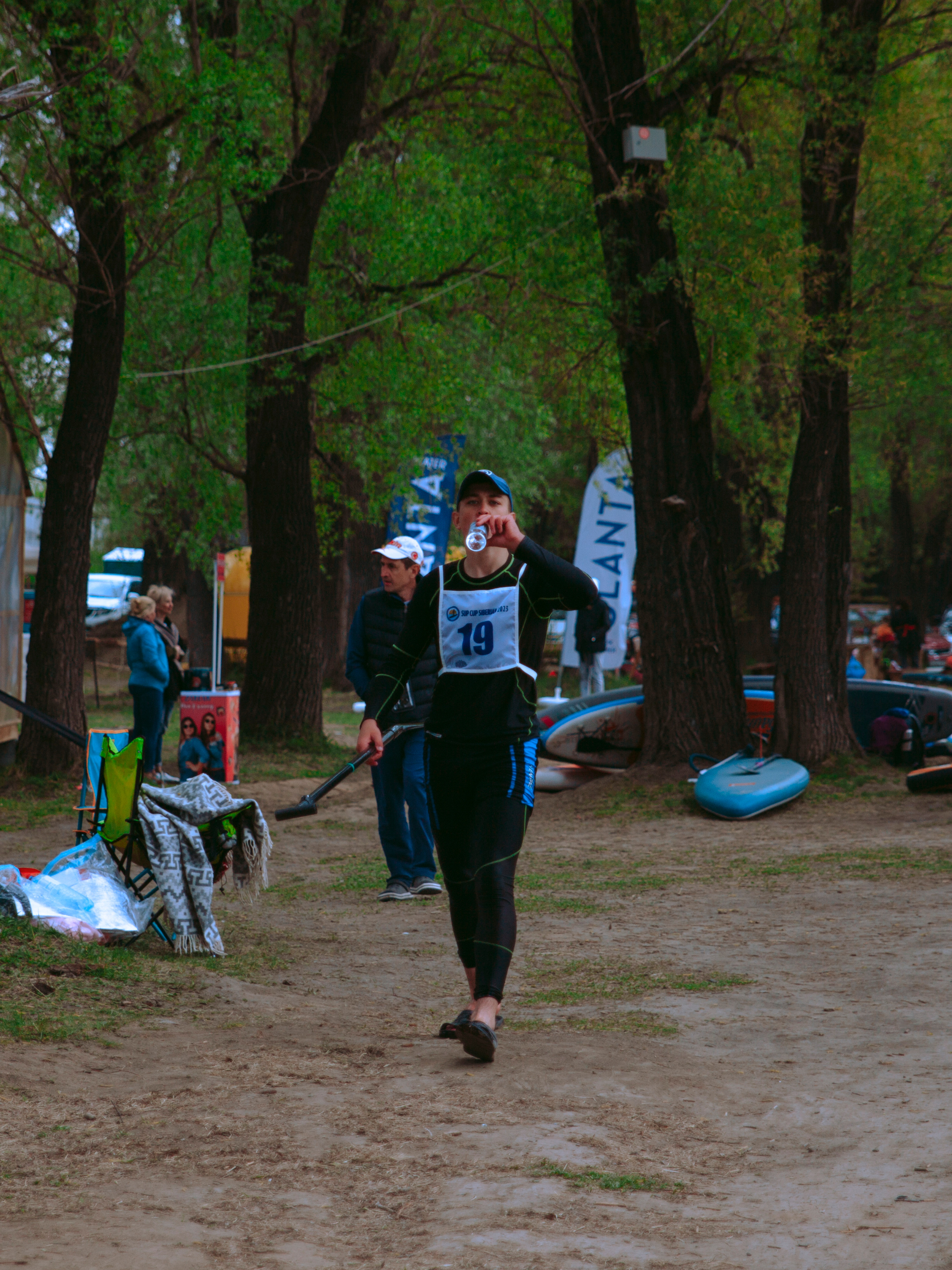 Спортсмен в гидрокостюме с номером на груди бежит и пьёт воду.