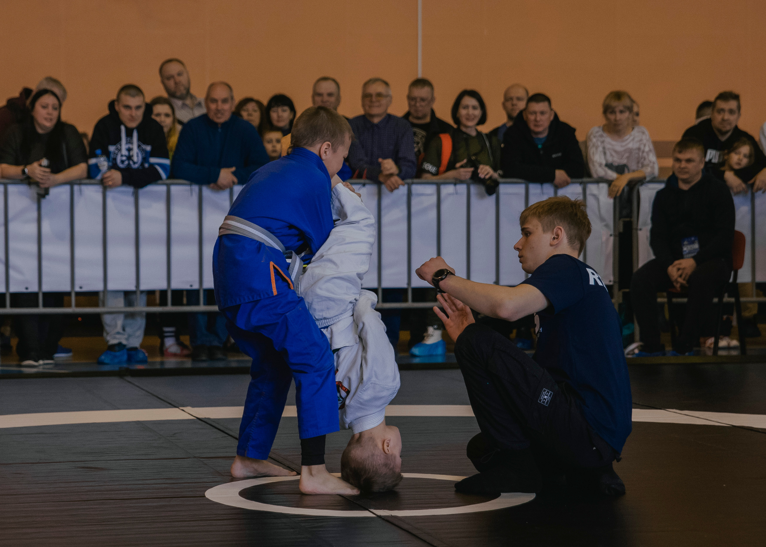 Один спортсмен держит другого вниз головой на детском турнире по джиу-джитсу.