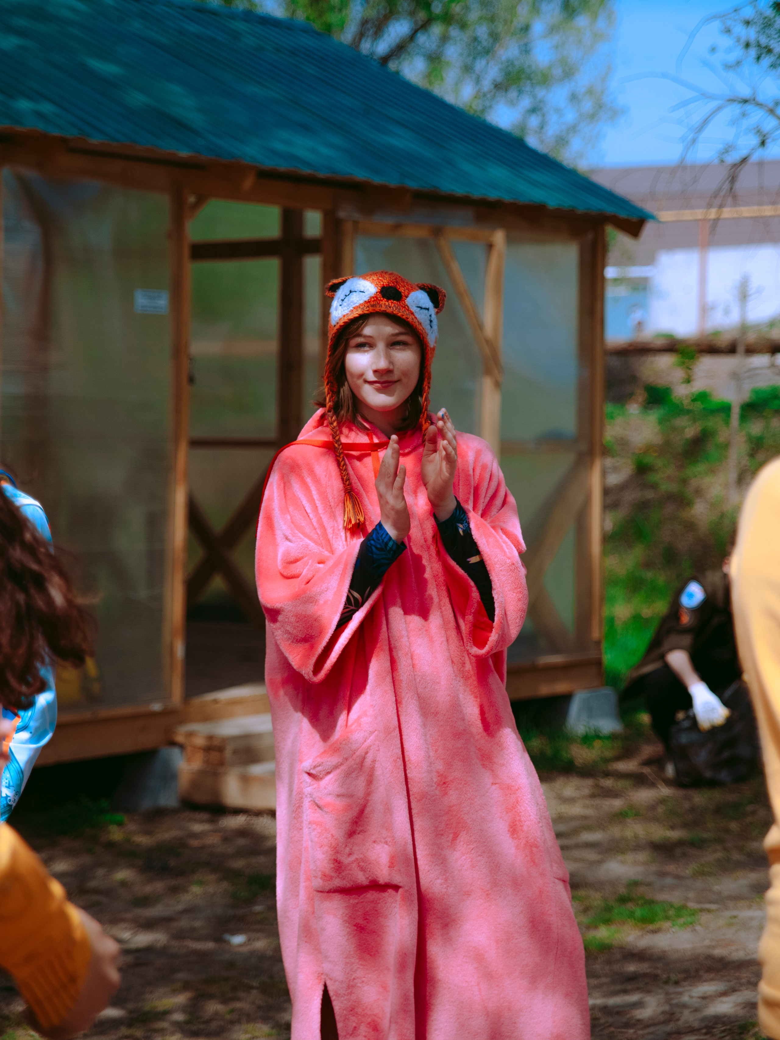 Девочка в розовом пончо и вязаной шапке в виде лисы радуется и хлопает в ладоши.