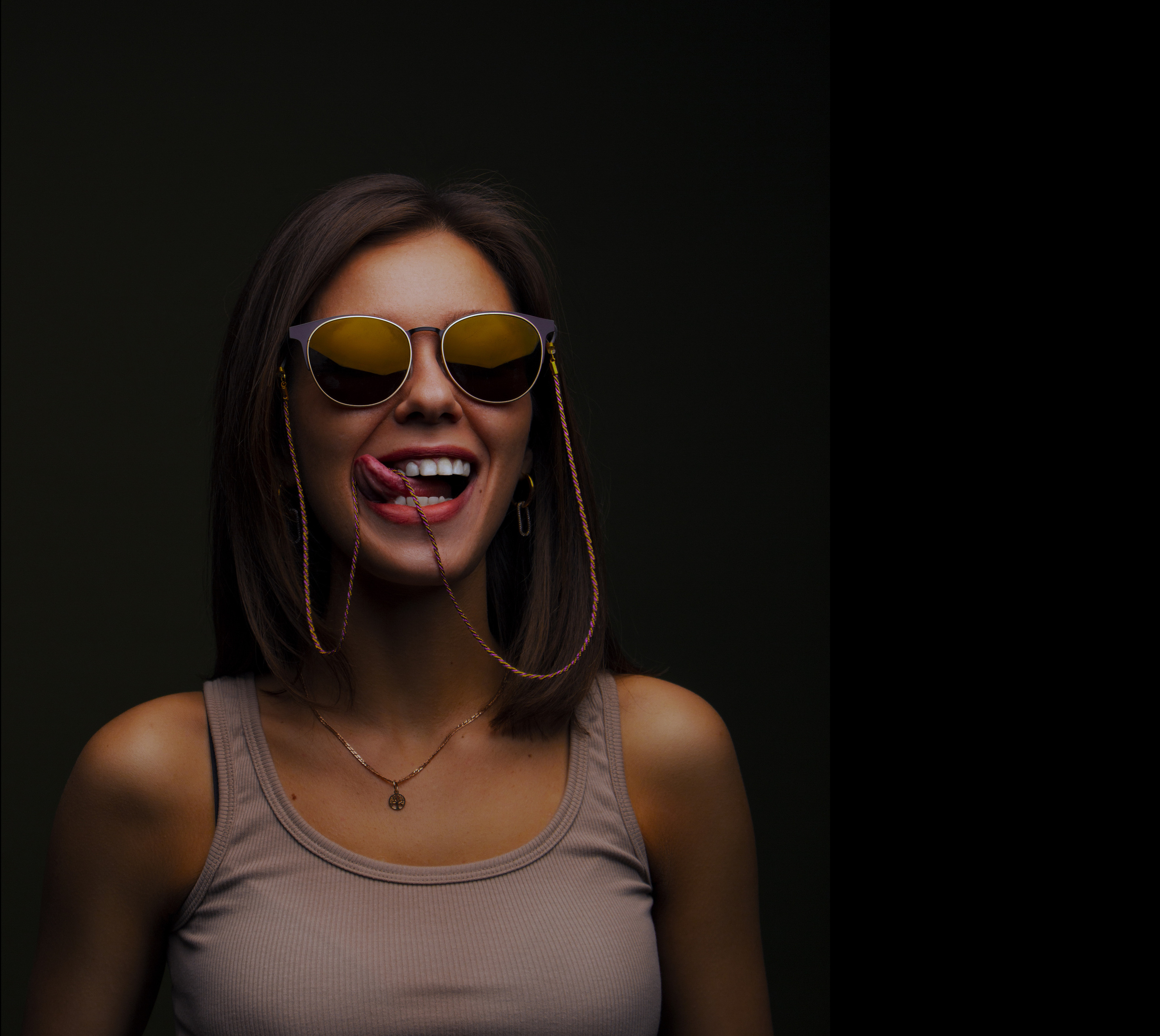 Девушка с белыми зубами в солнечных желтых очках с высунутыми языком облизывает губы.