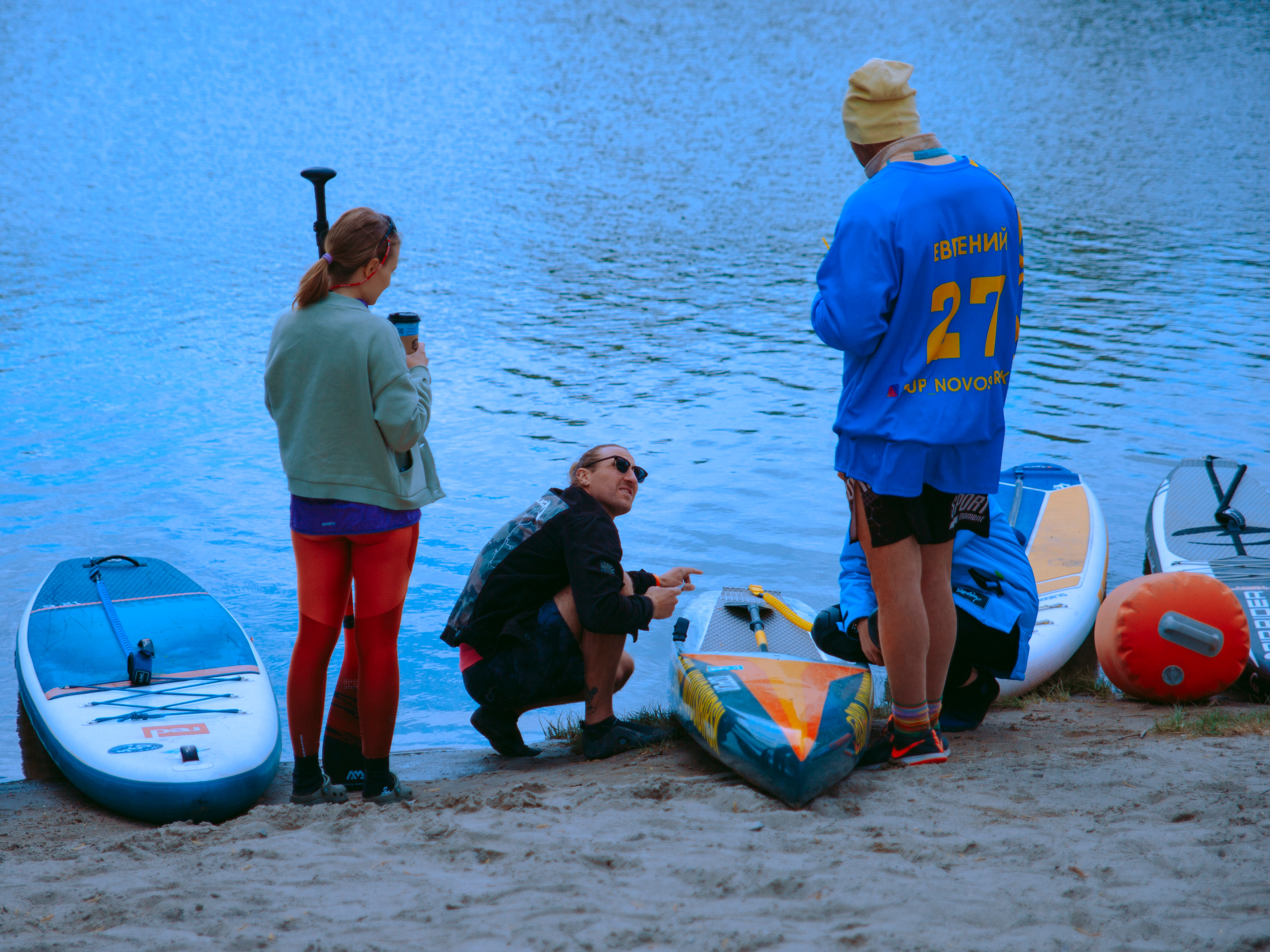Тренер по сап-сёрфингу силит на корточках у сапа на берегу.