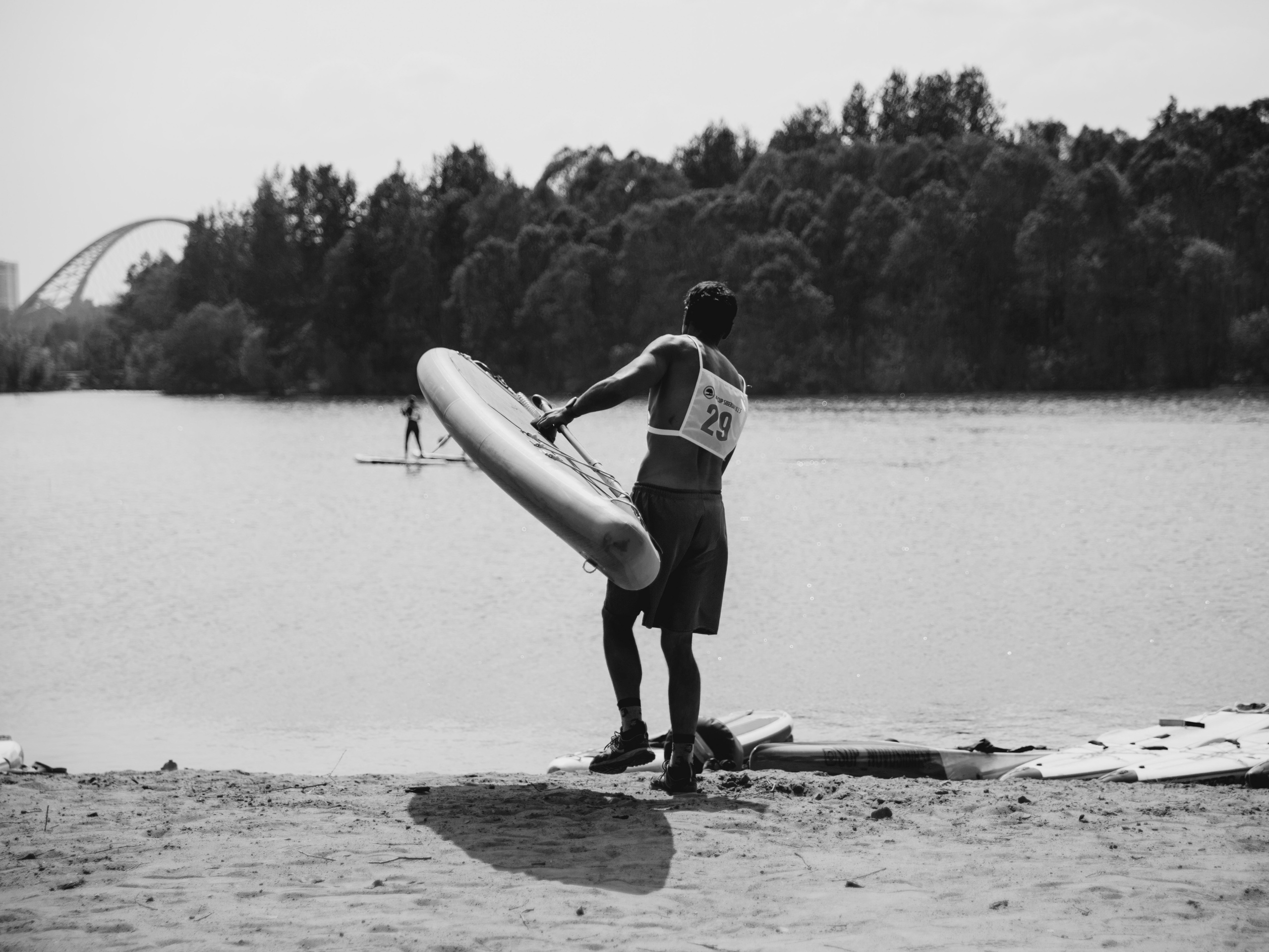 Мужчина с голым торсом на берегу несет сап в руках.