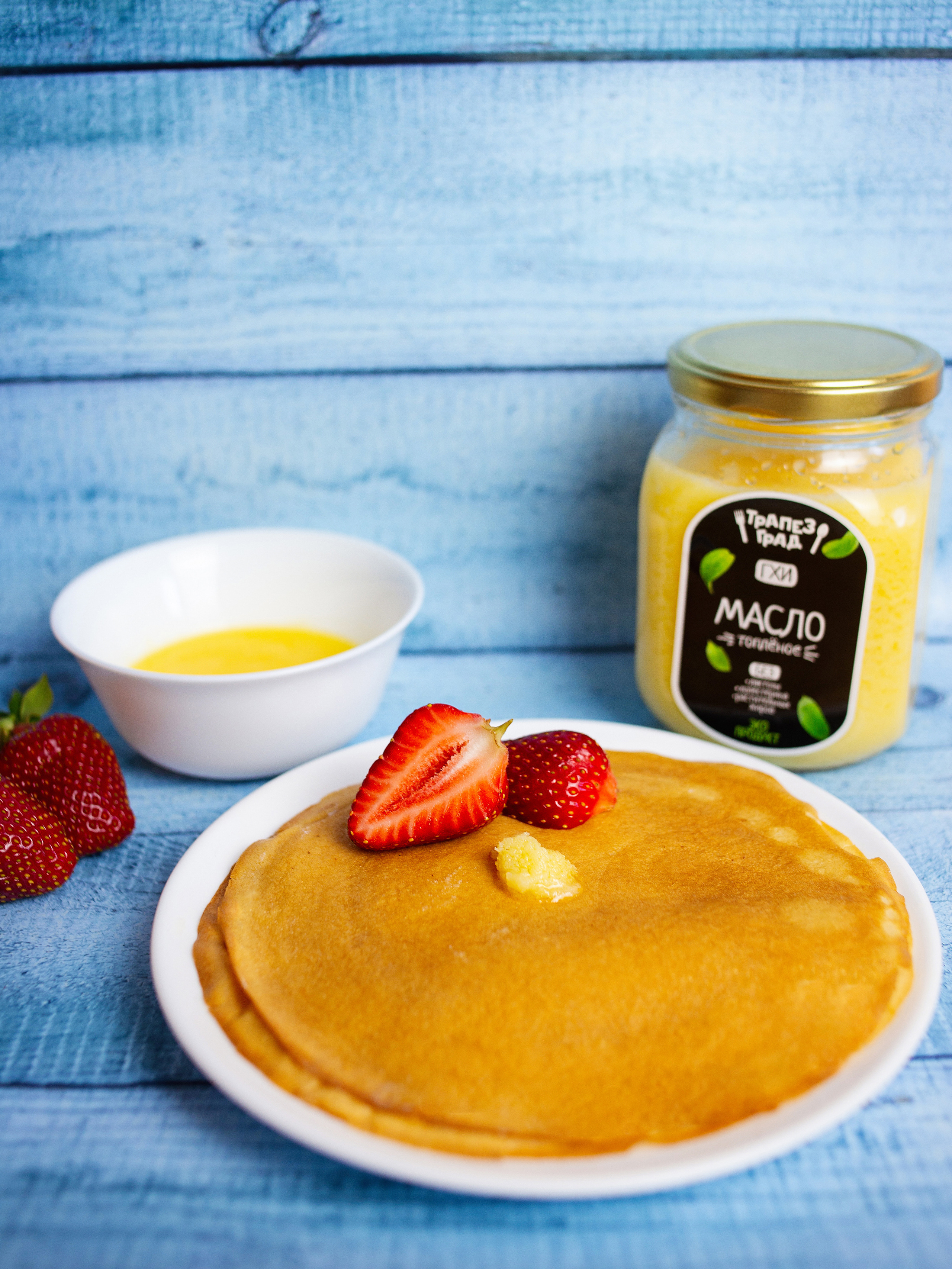 на столе  белая  тарелка   с блинчиками. она украшена клубникой. рядом стоит   топленное  масло